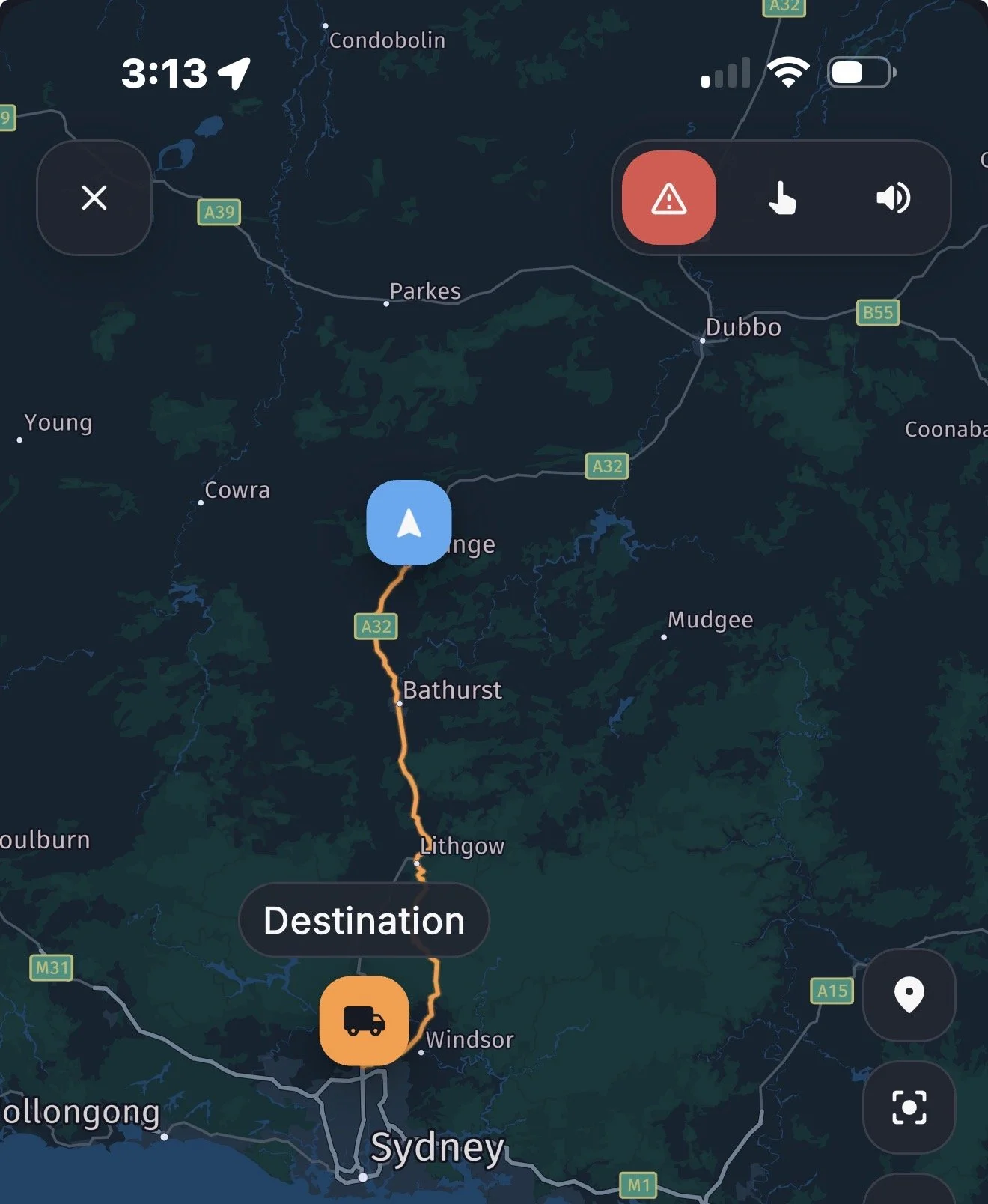 A GPS navigation screen showing a map of an area in New South Wales, Australia. The map shows roads, towns, and a route leading from Windsor to an unspecified destination. The time displayed is 3:13, and there are icons indicating network signal, Wi-Fi, and battery status. A red warning icon and a truck icon are also visible on the map.