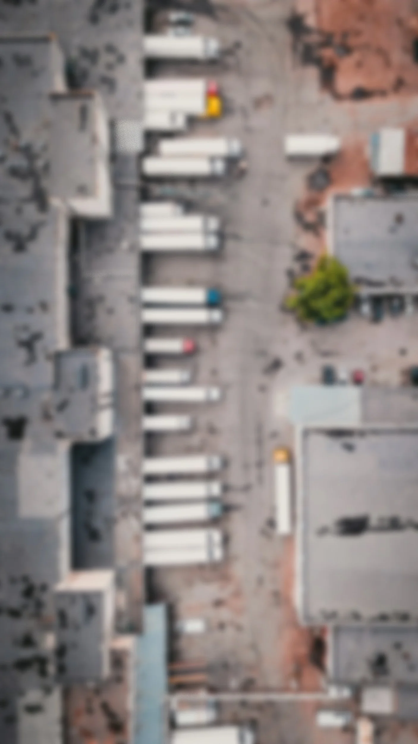 A blurry aerial view of a city street with parked cars, buildings, and a tree.