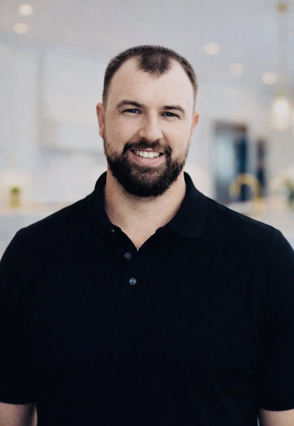 A smiling man with short brown hair, a beard, wearing a black polo shirt, standing indoors with a blurred background.
