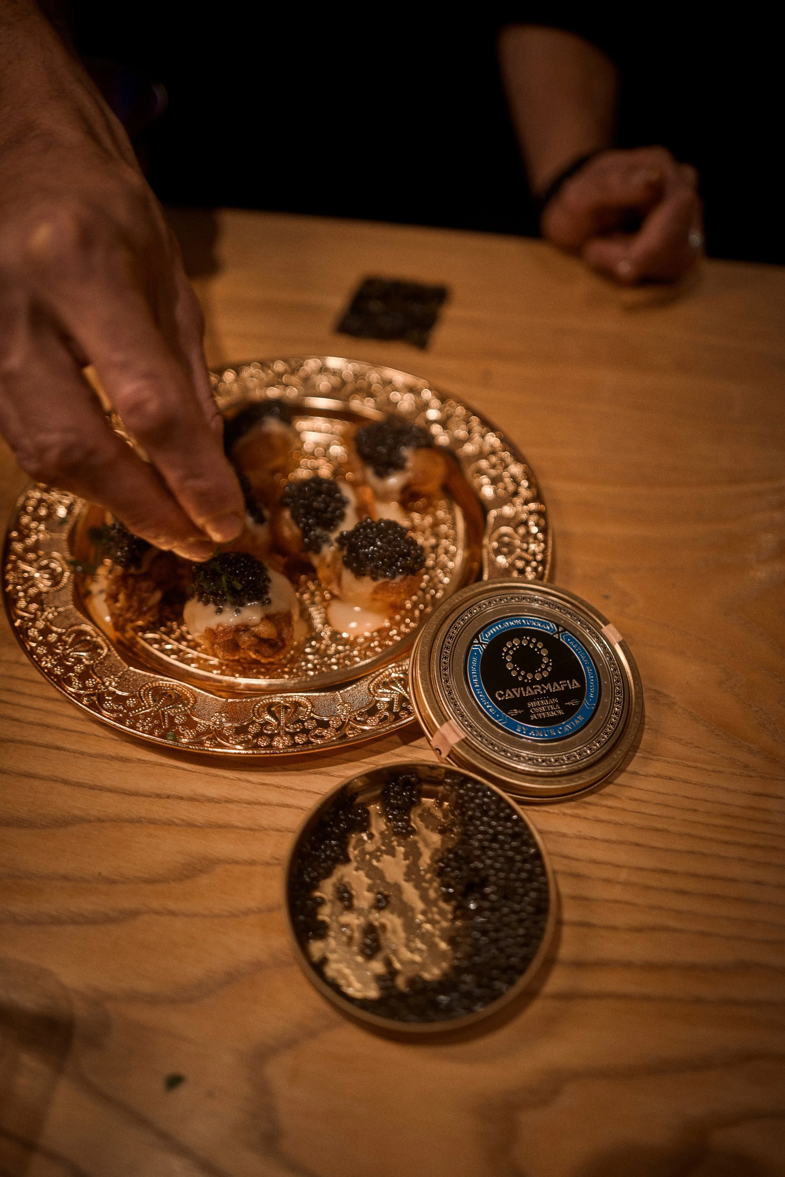 Person handling small appetizers topped with black caviar on an ornate silver plate, with a can of caviar on a wooden table.
