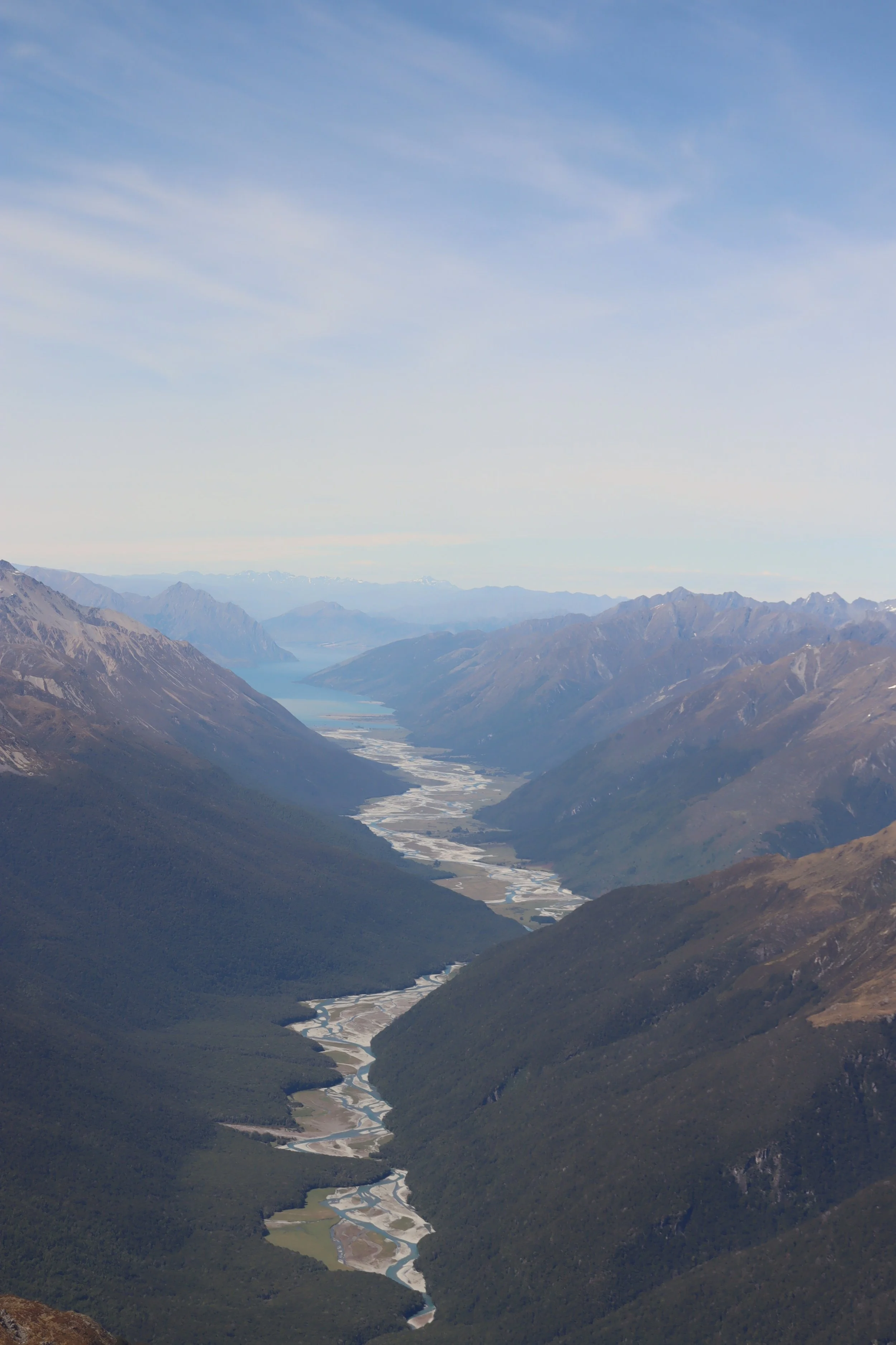 Aerial view of a winding river flowing through a mountainous valley with snow-capped peaks in the background under a blue sky with wispy clouds.