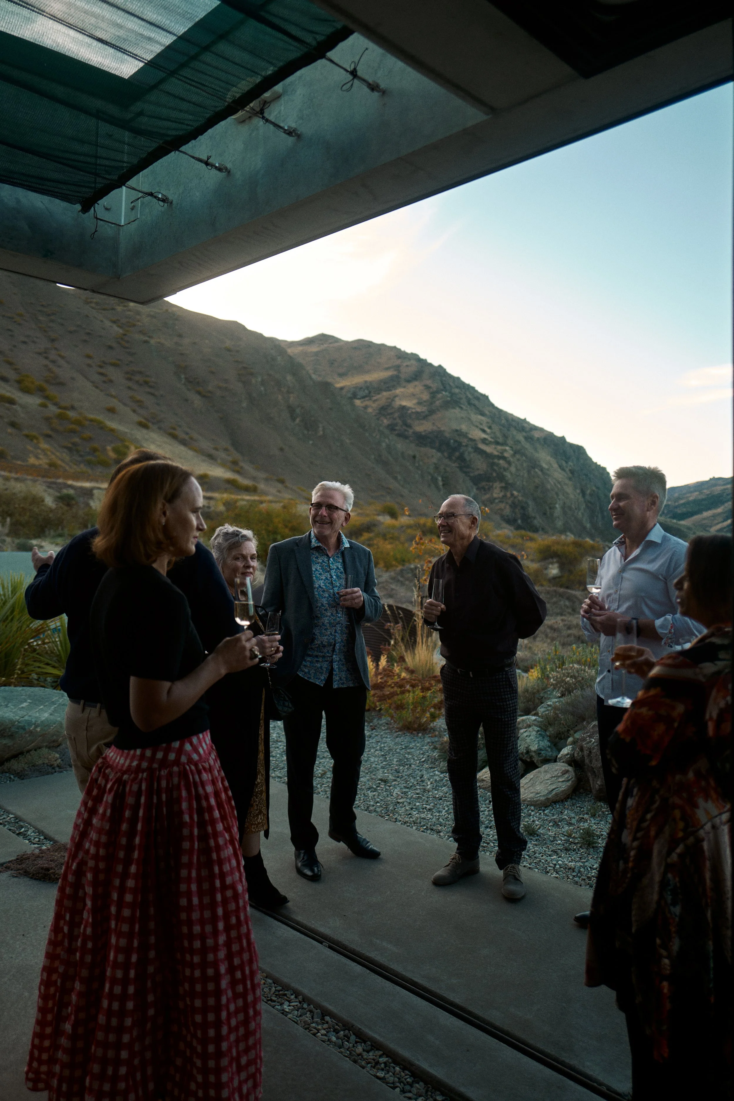 A group of seven people standing outdoors on a patio, enjoying drinks and conversing, with mountains and a sunset in the background.