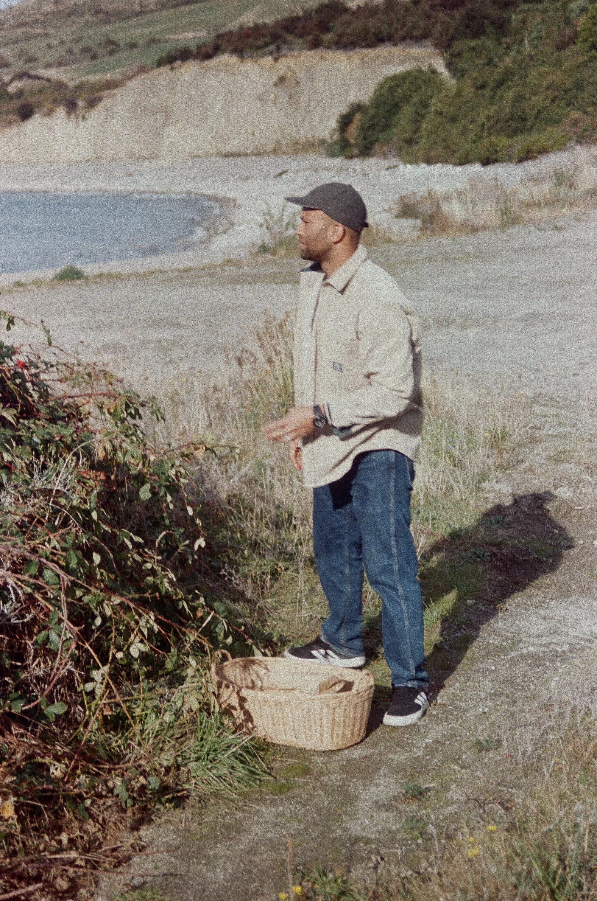 A man stands outdoors near the coast, holding a branch or bush, with a basket on the ground in front of him, wearing a light jacket, jeans, and a dark cap, on a sandy path with grass and bushes around.