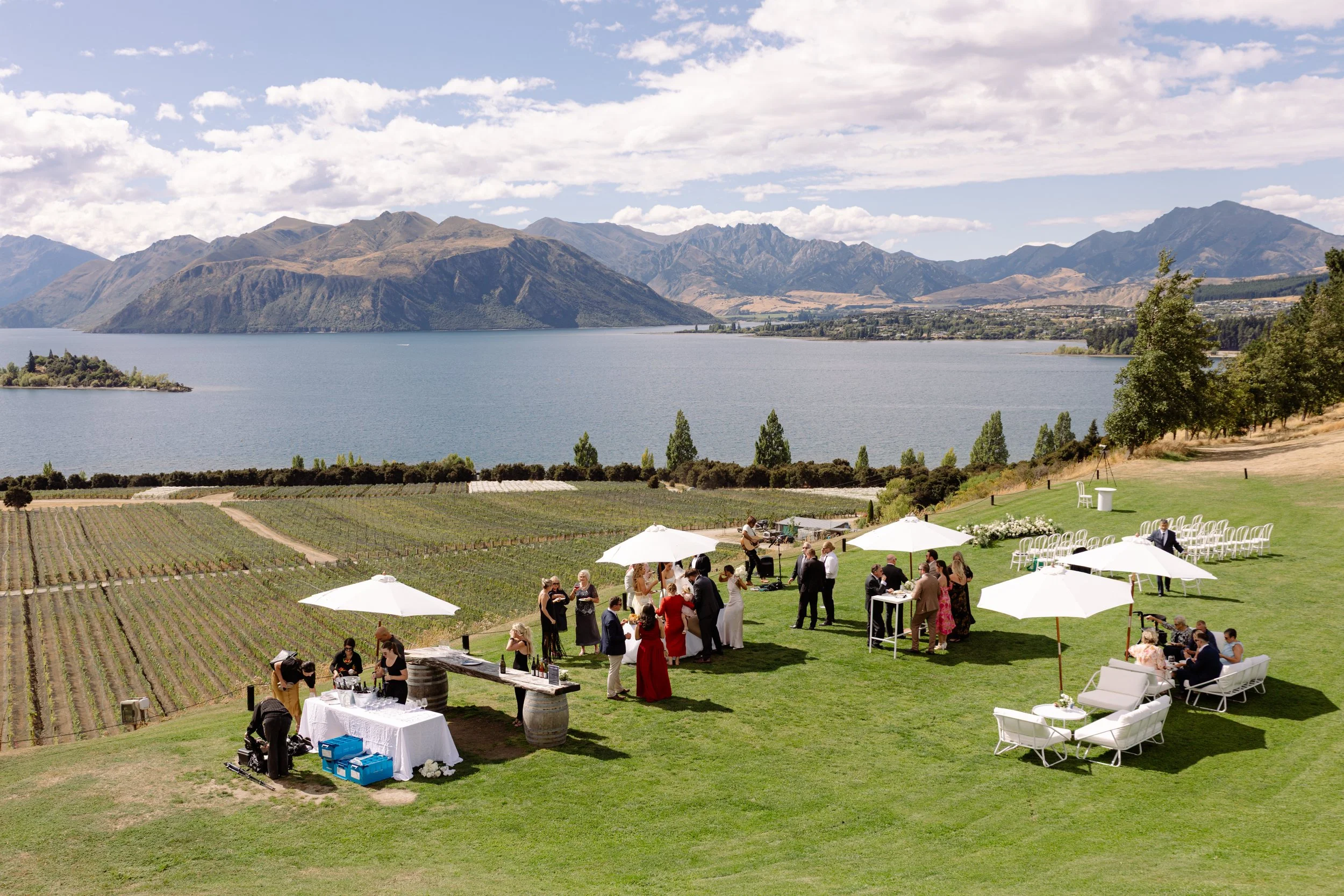 People at an outdoor celebration or wedding on a grassy hillside with a vineyard in the foreground, overlooking a lake surrounded by mountains.