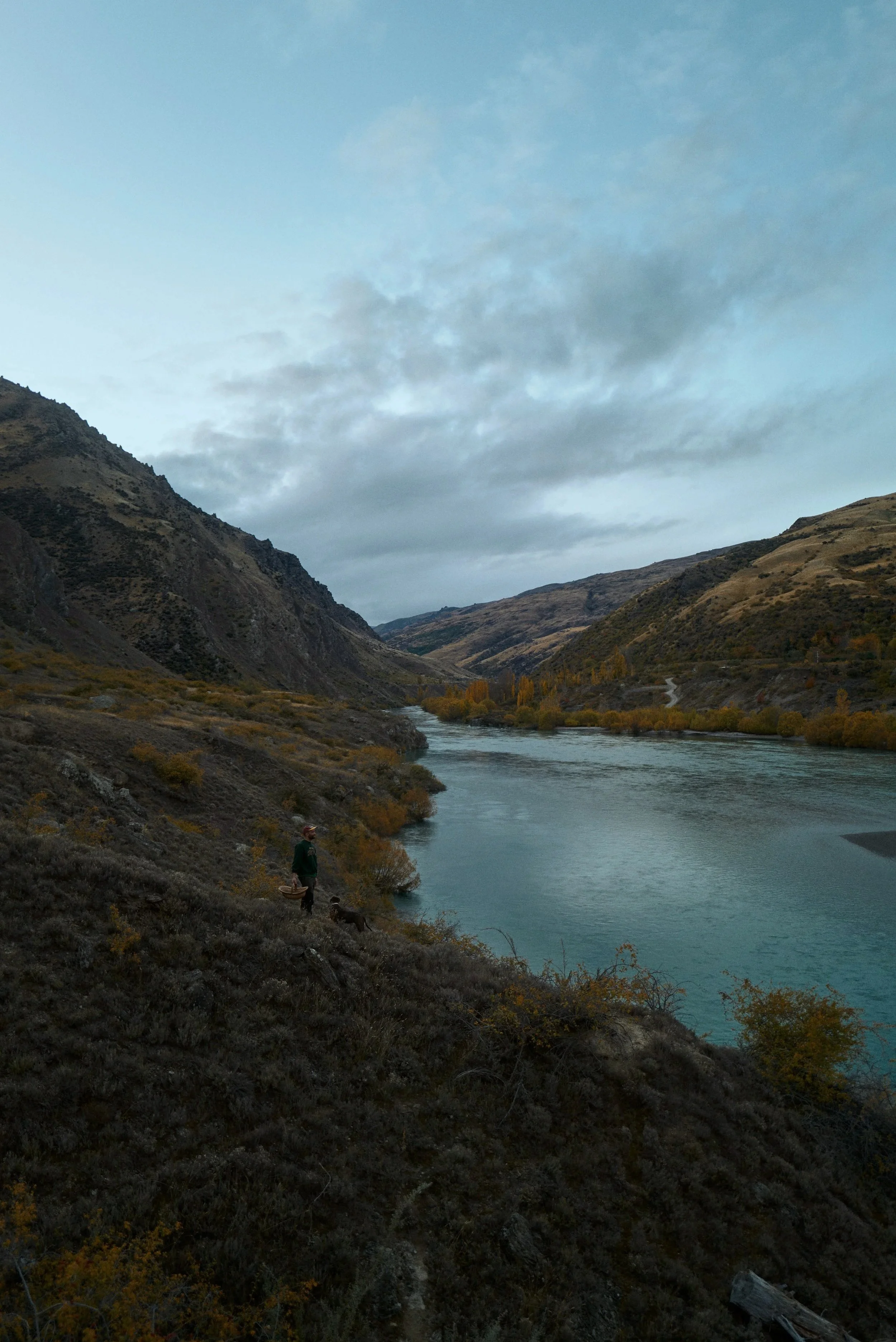 Person walking along a hillside by a river, surrounded by mountains under a cloudy sky.