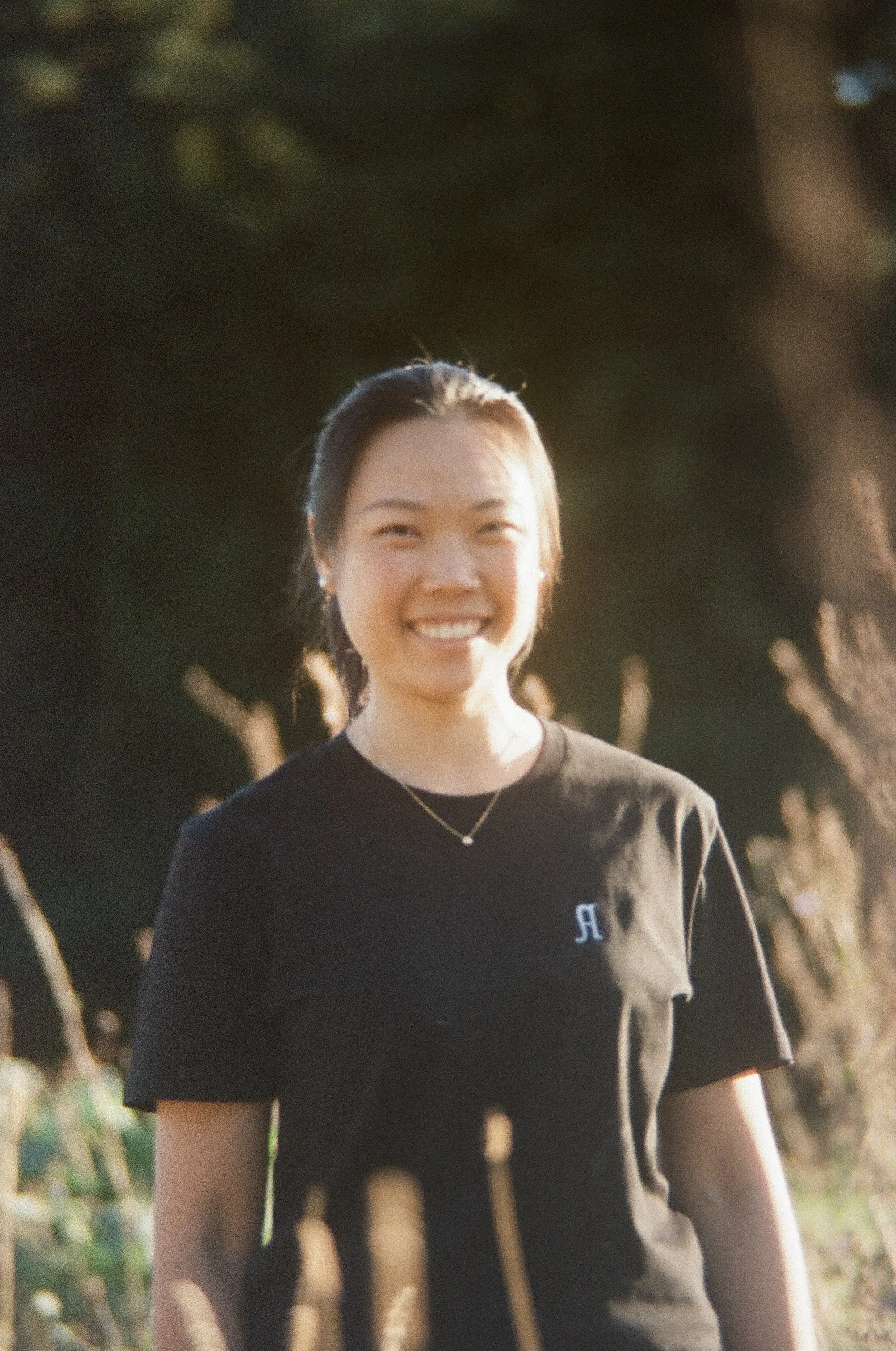 A smiling woman with black hair tied back, wearing a black t-shirt with a small Russian letter 'Я' on it, outdoors with blurred trees in the background.