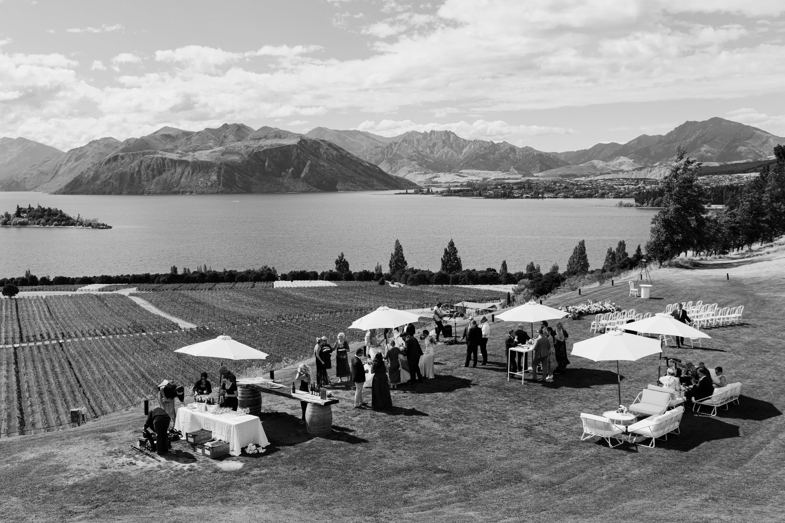 A black and white photo of an outdoor wedding reception on a grassy lawn overlooking a lake with mountains in the background. Guests are gathered around tables with umbrellas, with some seated and others standing. There is a small bar setup with bottles, and a guitarist performing. Rows of white chairs are arranged for guests, and trees line the edge of the lawn.