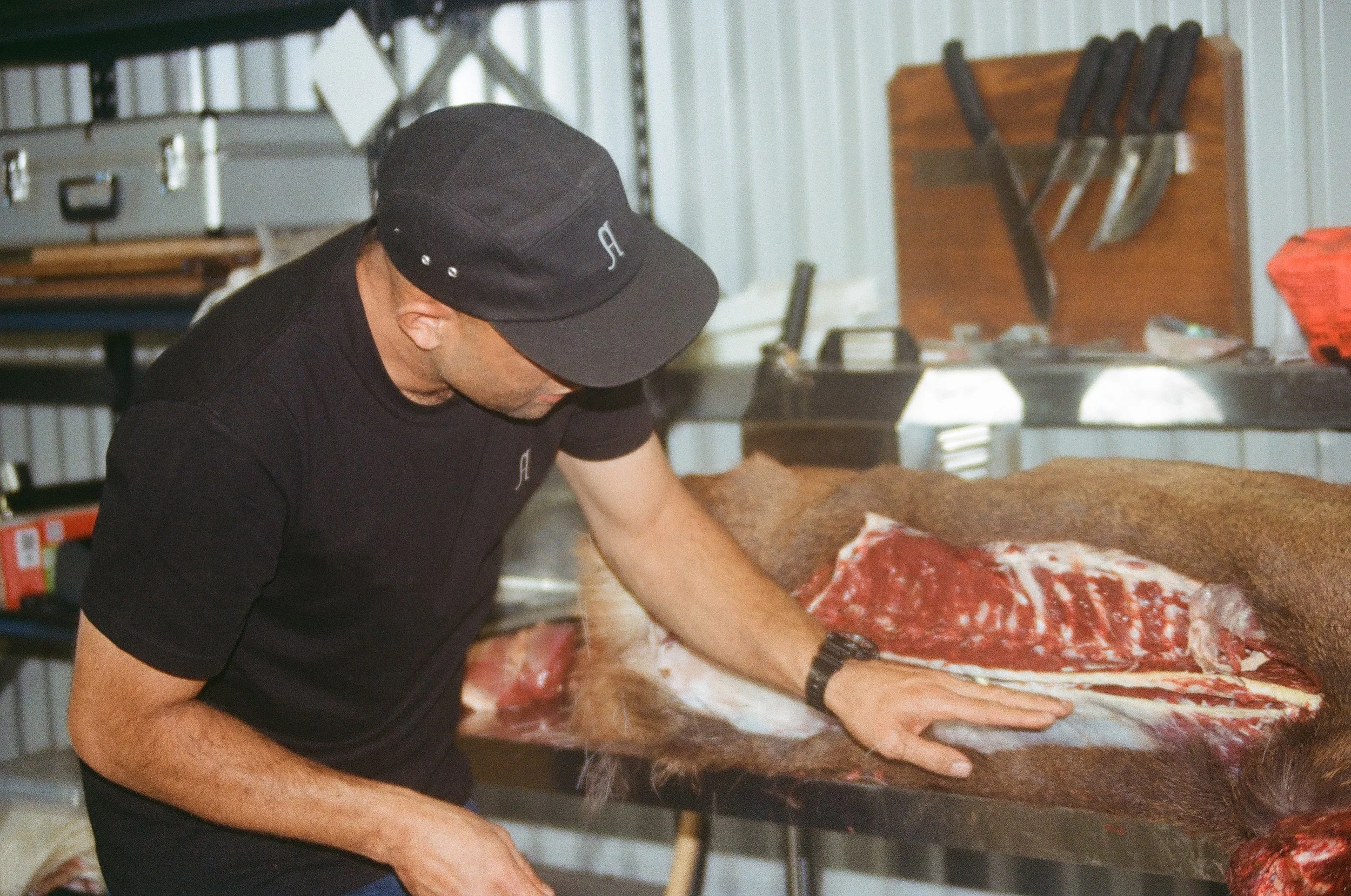 A man wearing a black cap and black T-shirt is processing a large animal carcass on a wooden work table inside a meat processing facility. The carcass is partially skinned, revealing muscle and bone, and has brown fur.