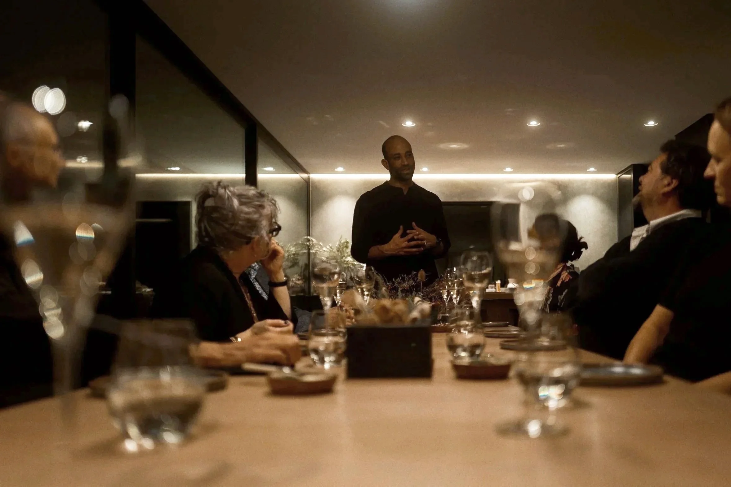 A group of people sitting around a dinner table listening to a speaker standing at the end of the table in a dimly lit restaurant.