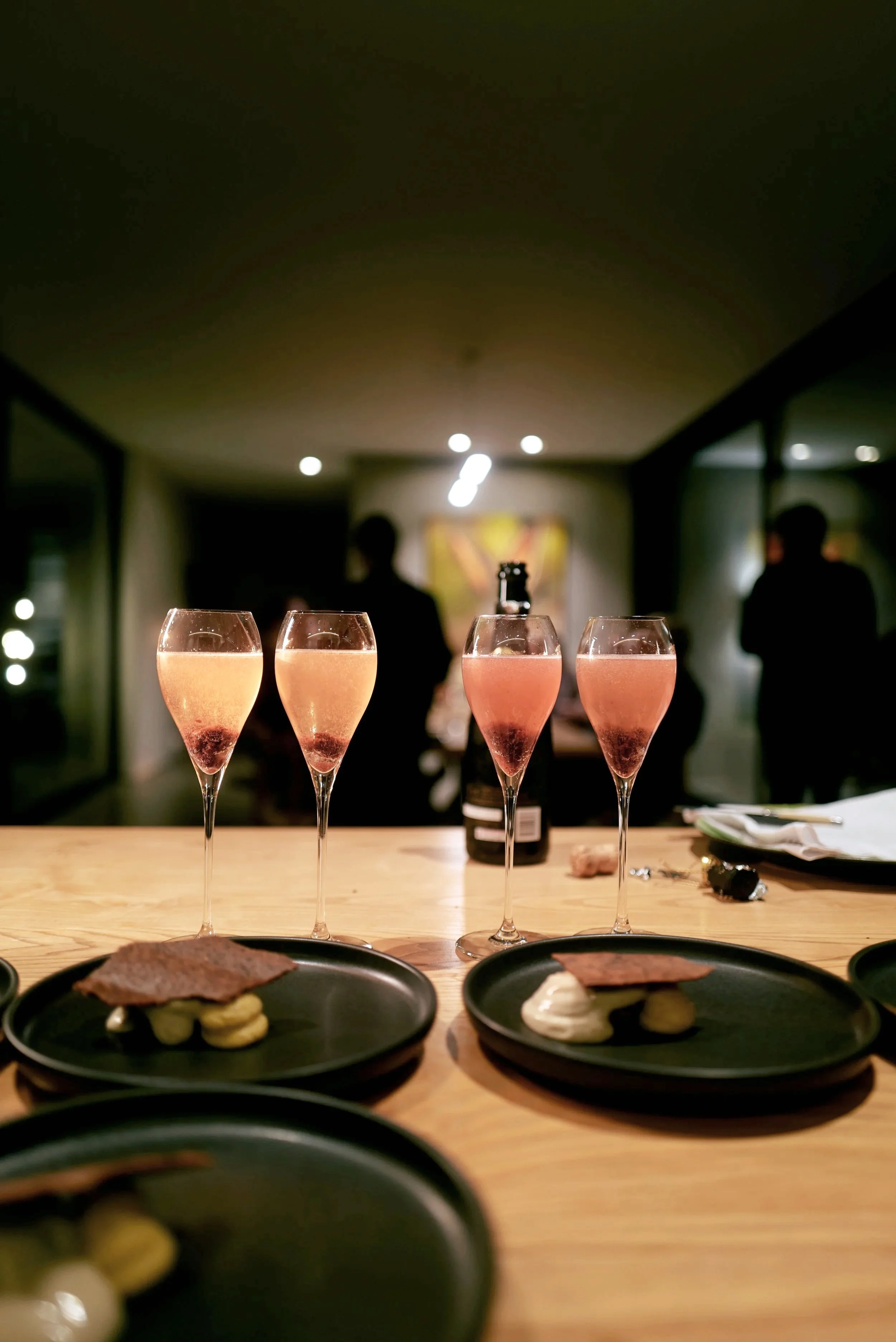 Four glasses of pink sparkling wine on a wooden table with plates of food, a bottle of sparkling wine, and a cork, in a dimly lit room with silhouettes of people in the background.