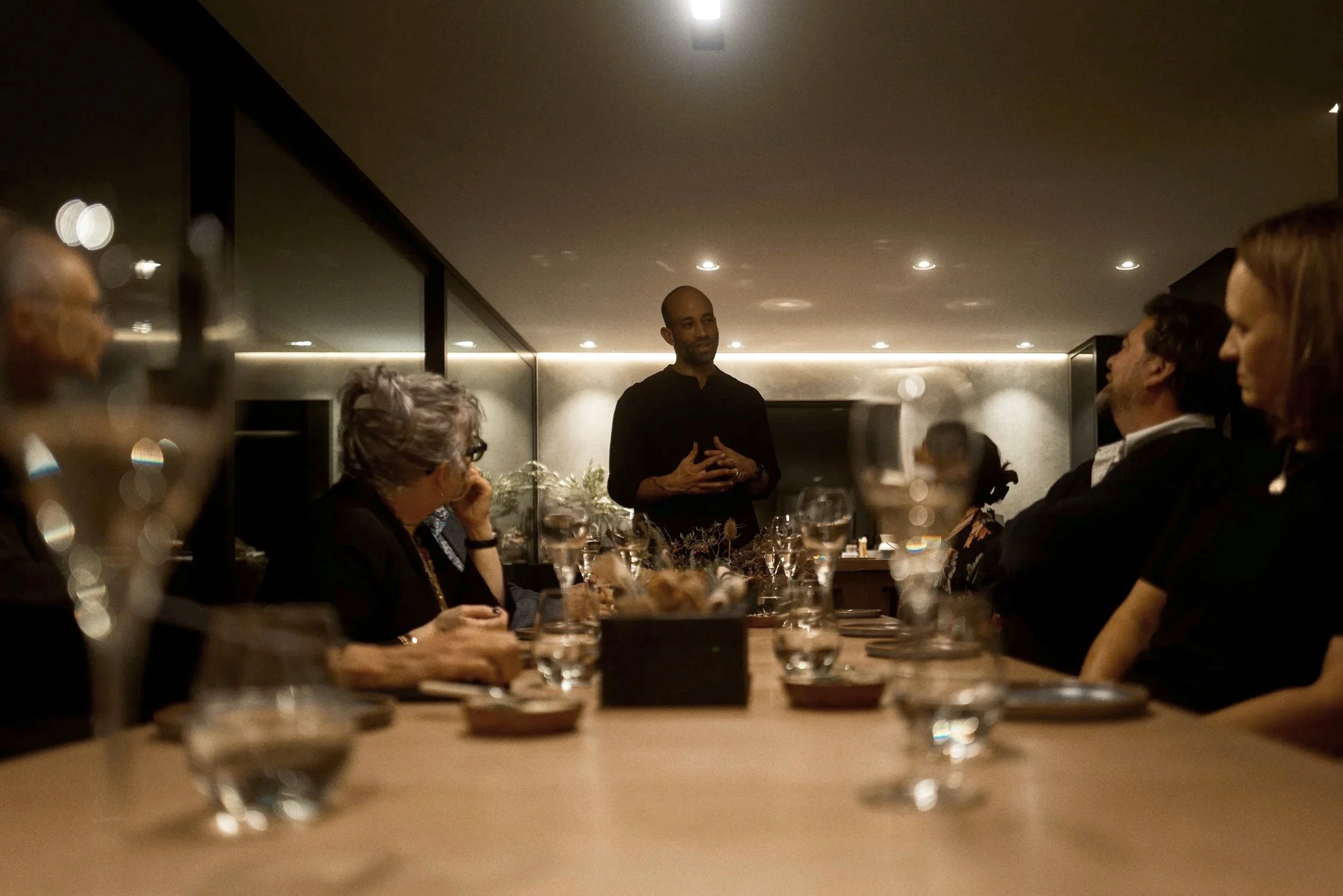 A group of people seated around a dining table listening to a man who is standing and speaking in a modern, dimly lit room.