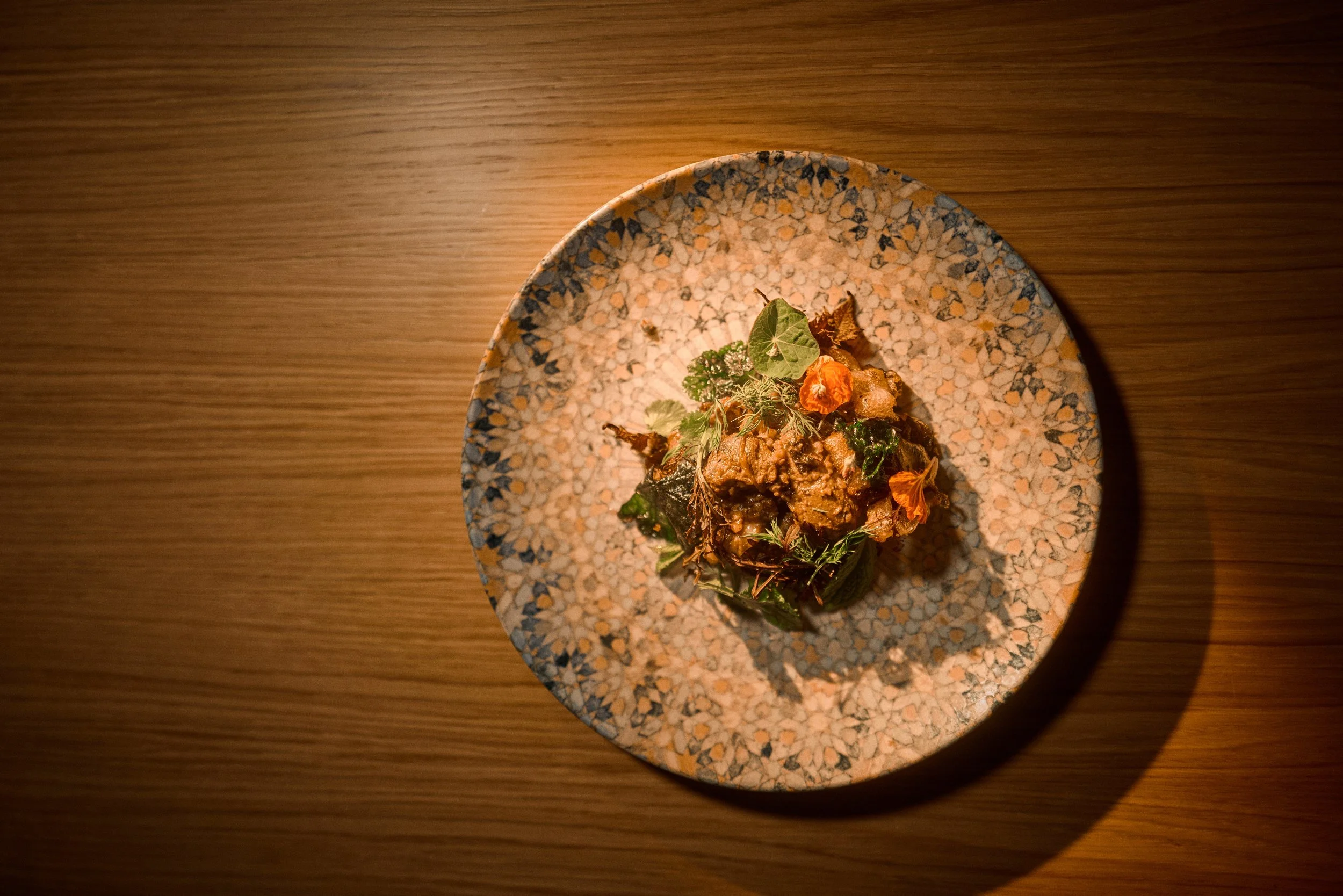 A dish of cooked meat garnished with herbs and edible flowers, served on a speckled ceramic plate, with warm lighting on a wooden surface.