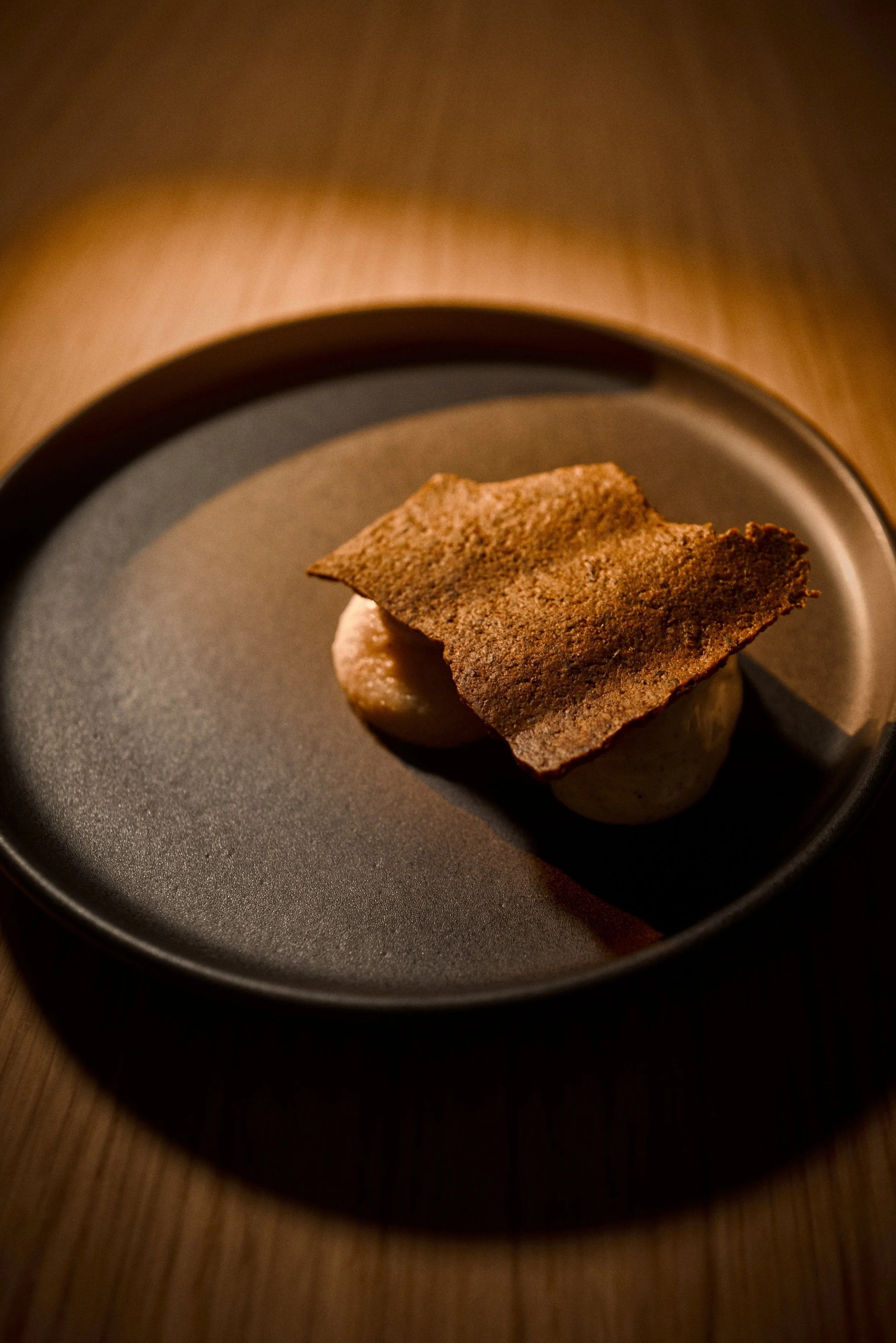 A plated dessert featuring a crispy, dark brown sheet on top of three round, white dollops on a black plate.