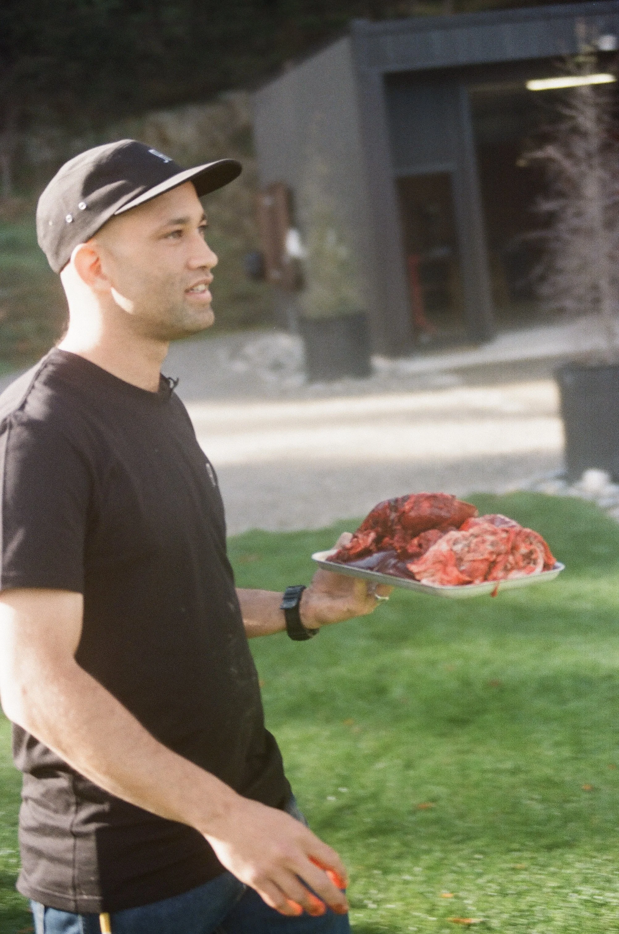 A man holds a plate with raw, red meat outdoors, with a building and grassy area in the background.