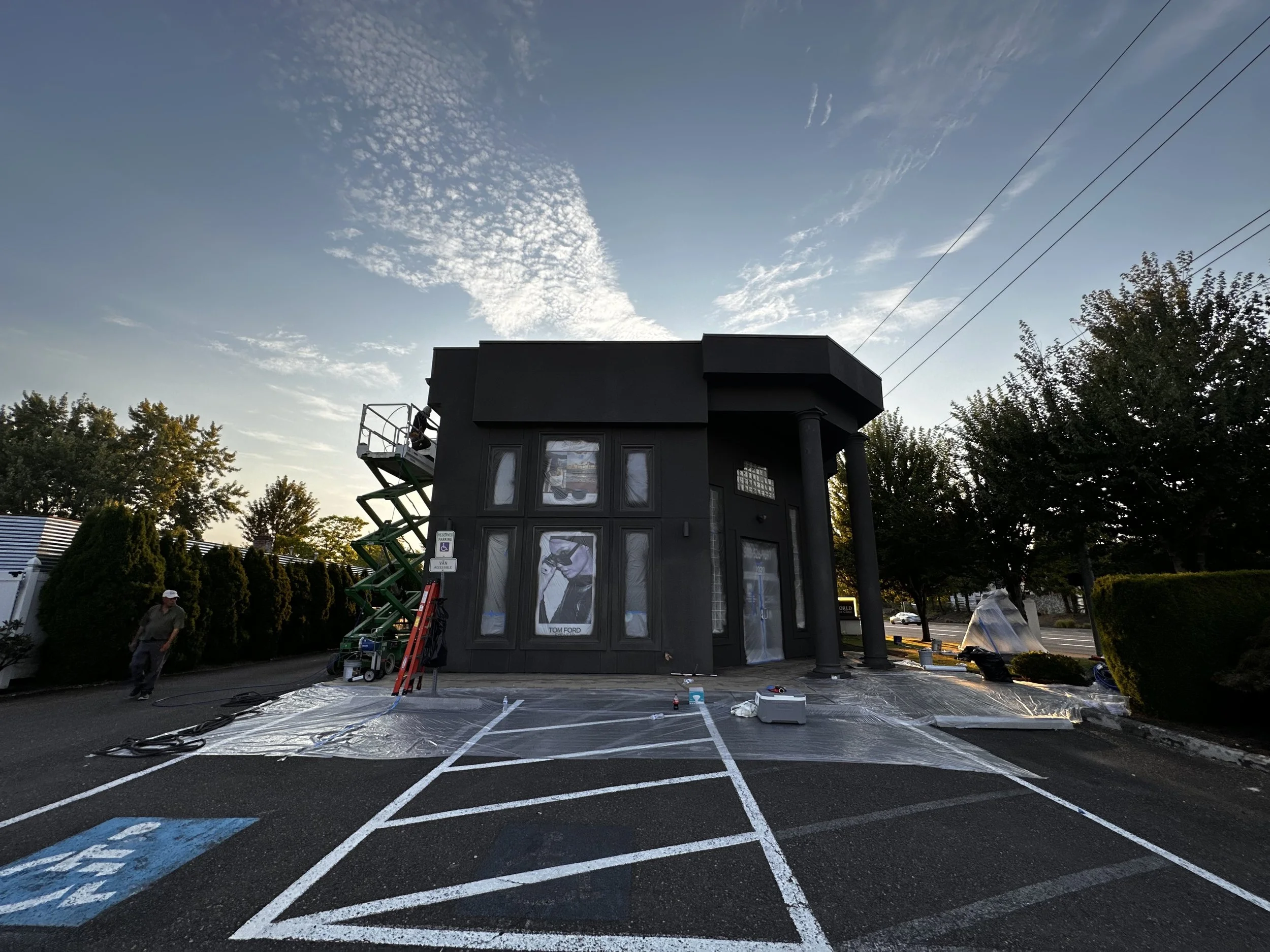 A black building under construction with plastic covering the ground outside, and a green scissor lift on the left side. There are trees and a parking lot with painted lines and handicap spots in the foreground.