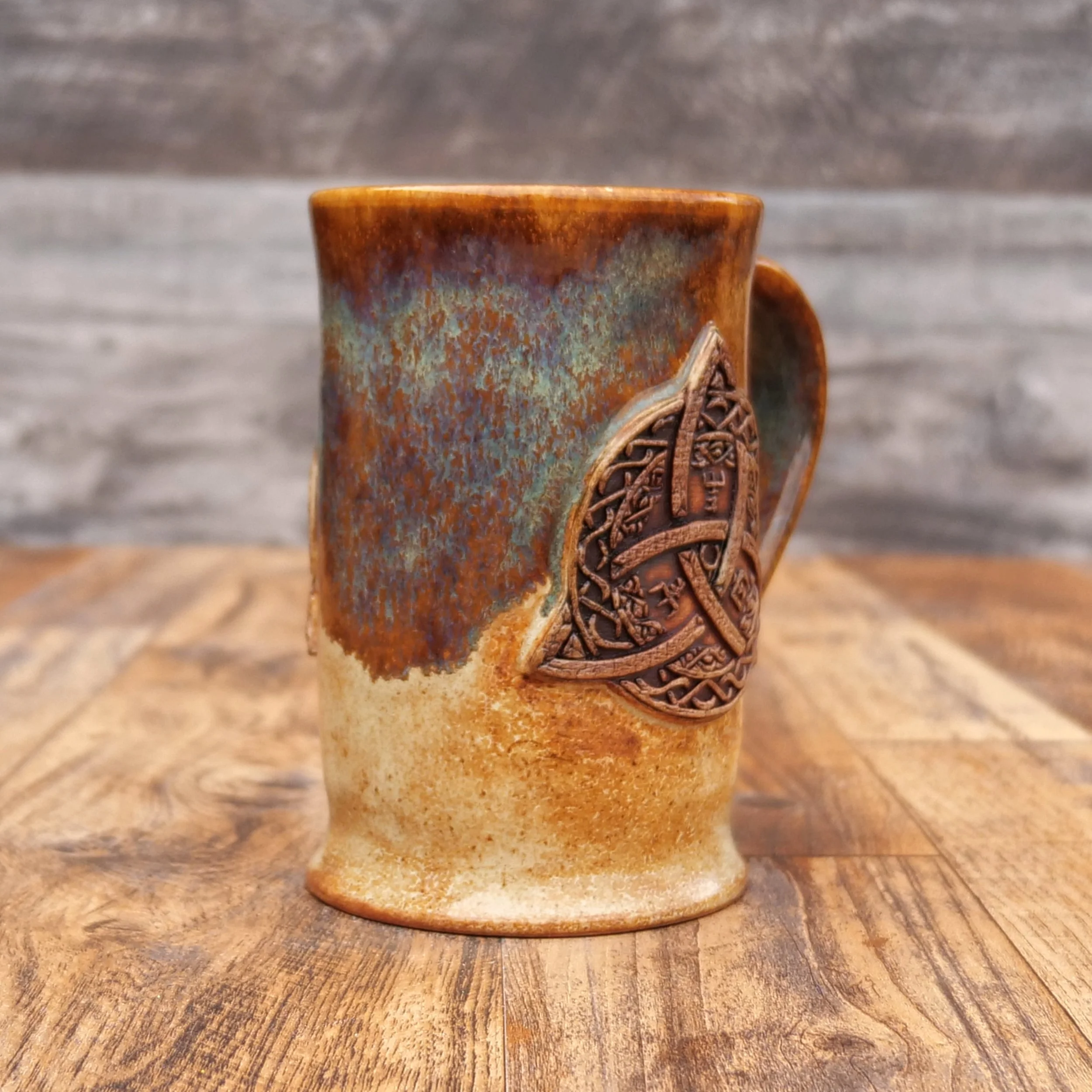 Handmade Trinity Knot Mug by Elderkiln Ceramics featuring a sculpted Celtic knot design and layered earthy brown and mineral-toned stoneware glazes.