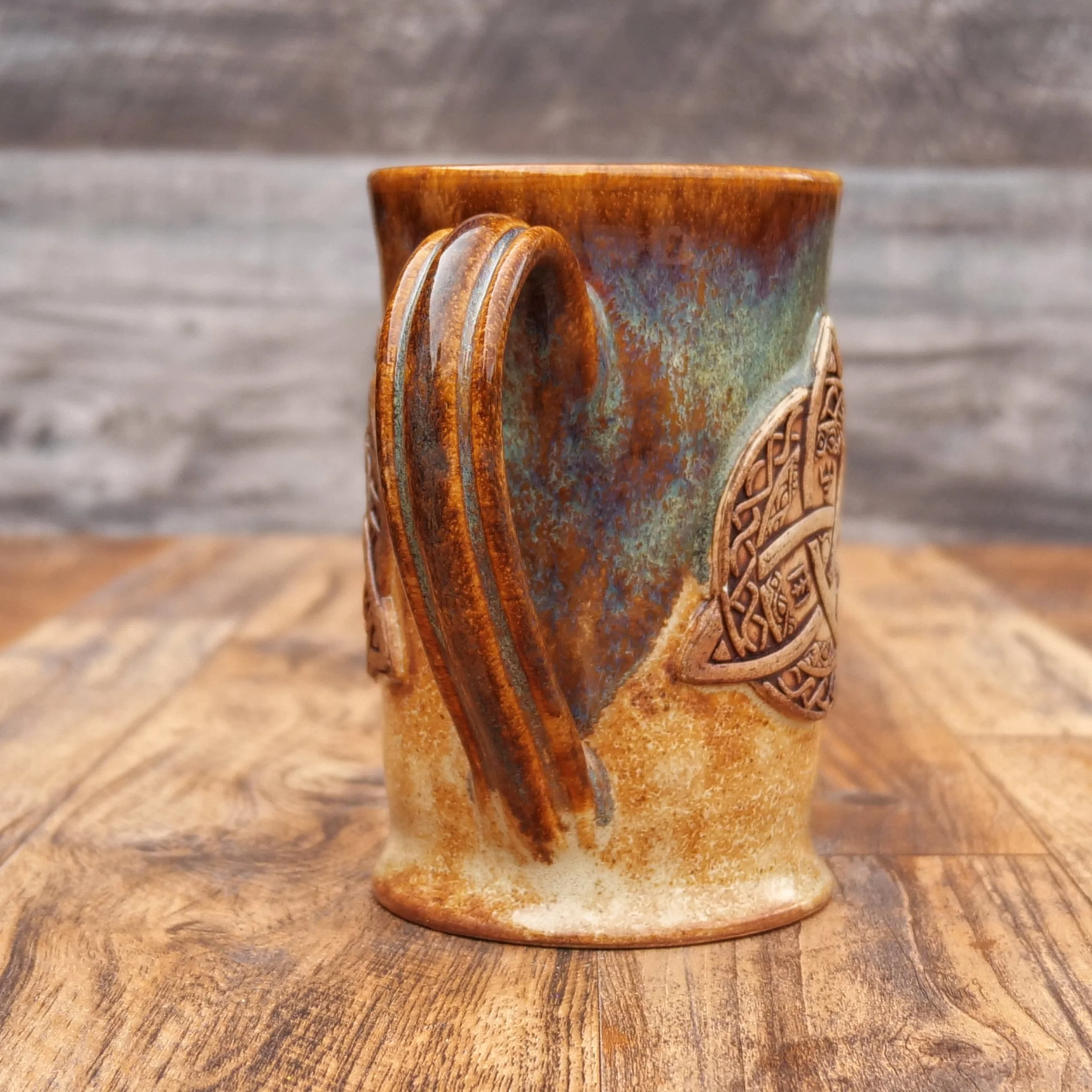 Handmade Trinity Knot Mug by Elderkiln Ceramics featuring a sculpted Celtic knot design and layered earthy brown and mineral-toned stoneware glazes.