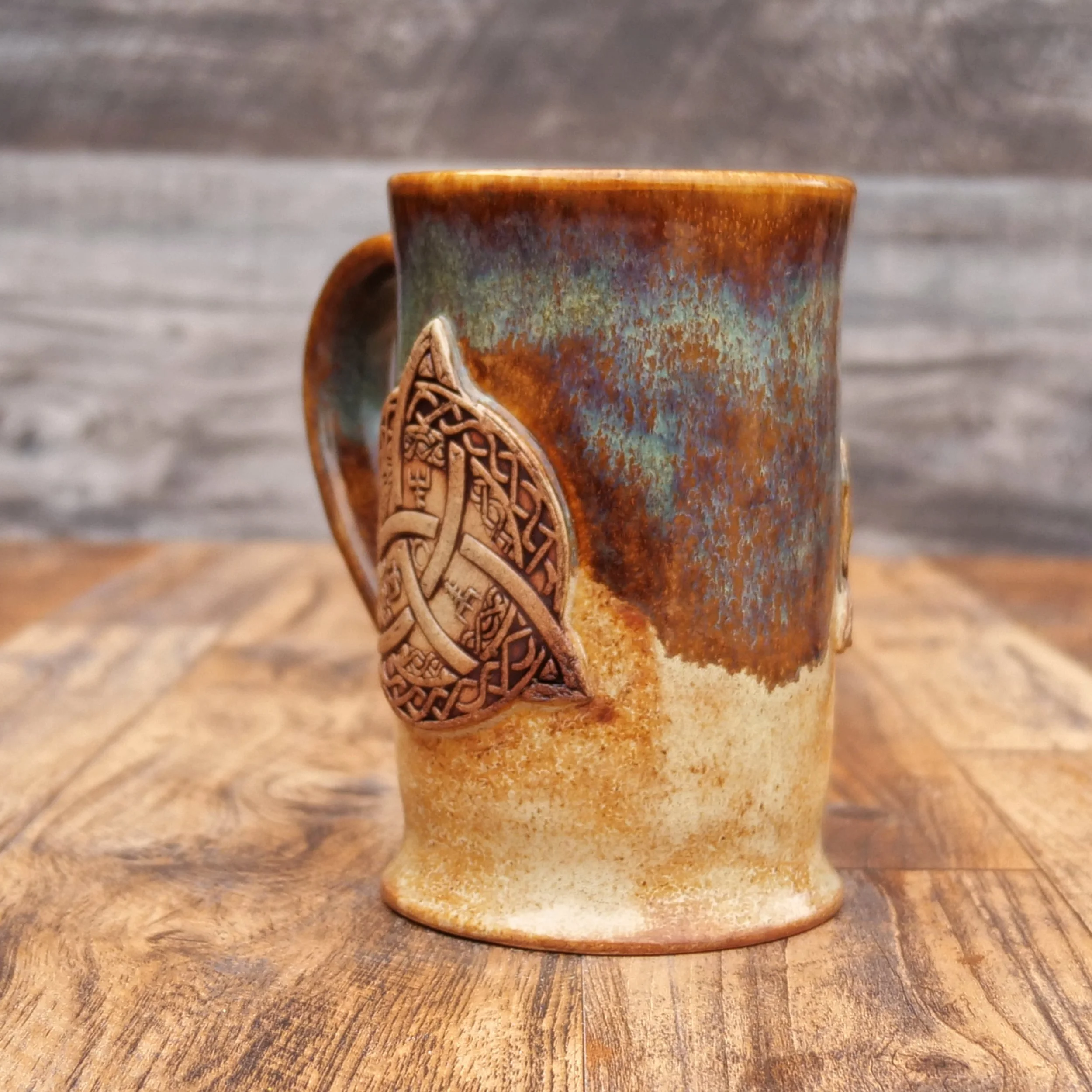 Handmade Trinity Knot Mug by Elderkiln Ceramics featuring a sculpted Celtic knot design and layered earthy brown and mineral-toned stoneware glazes.
