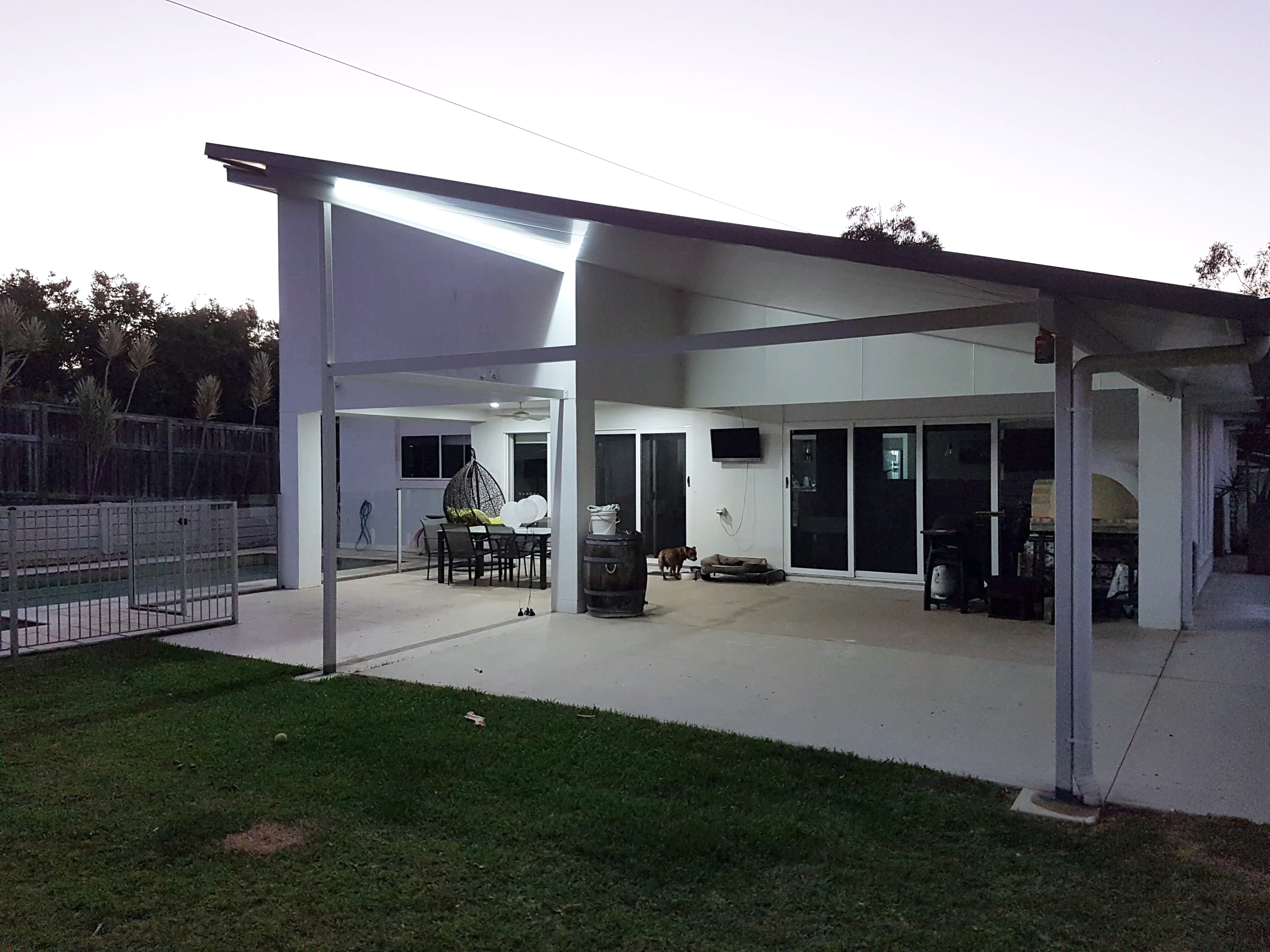 A backyard patio area at dusk with outdoor furniture, a hanging chair, a barbecue grill, and a dog near a small mattress. The patio is covered with a roof, and the house has large sliding glass doors. There is a patio table with chairs and various outdoor decor, with trees and a fence in the background.