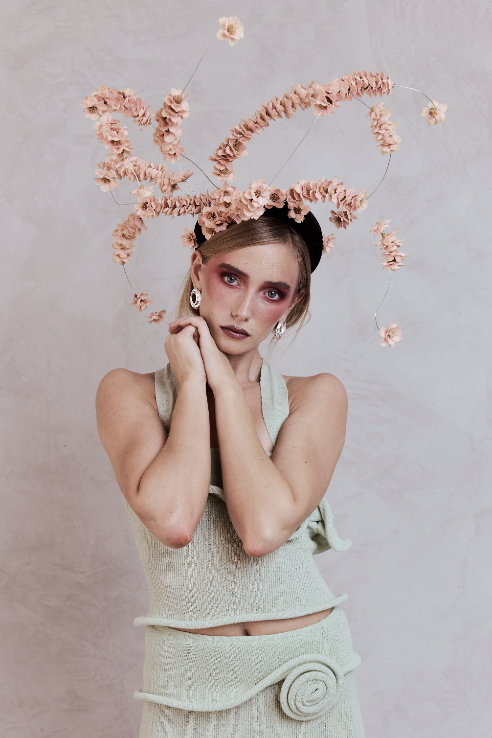 A woman with light skin, blue eyes, and styled blonde hair, wearing a beige sleeveless top and matching skirt with a spiral detail, poses with her hands near her face. She is accessorized with large silver earrings and has dramatic pink eye makeup an