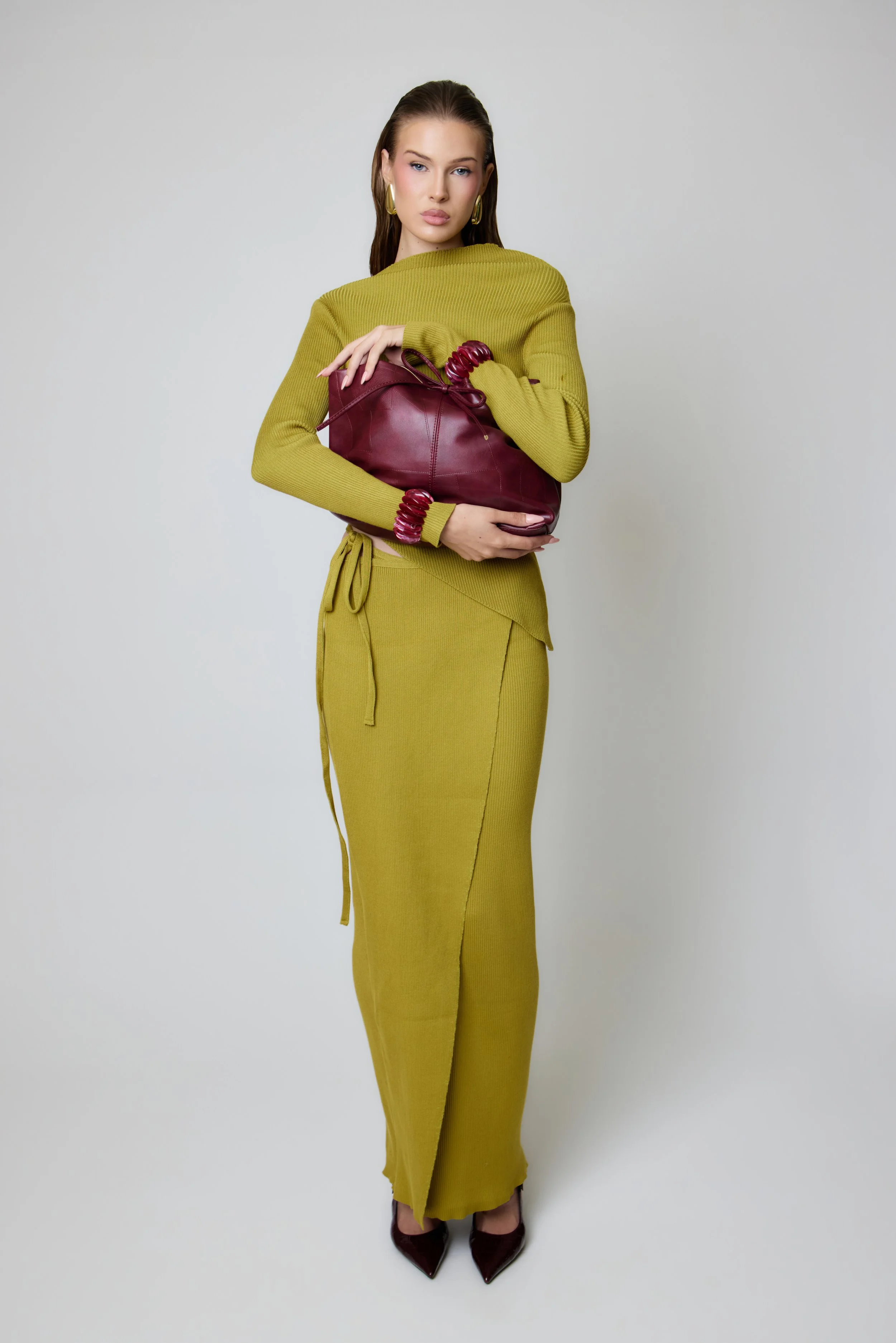 A woman in a green long-sleeve dress, holding a maroon handbag, standing against a plain light gray background.