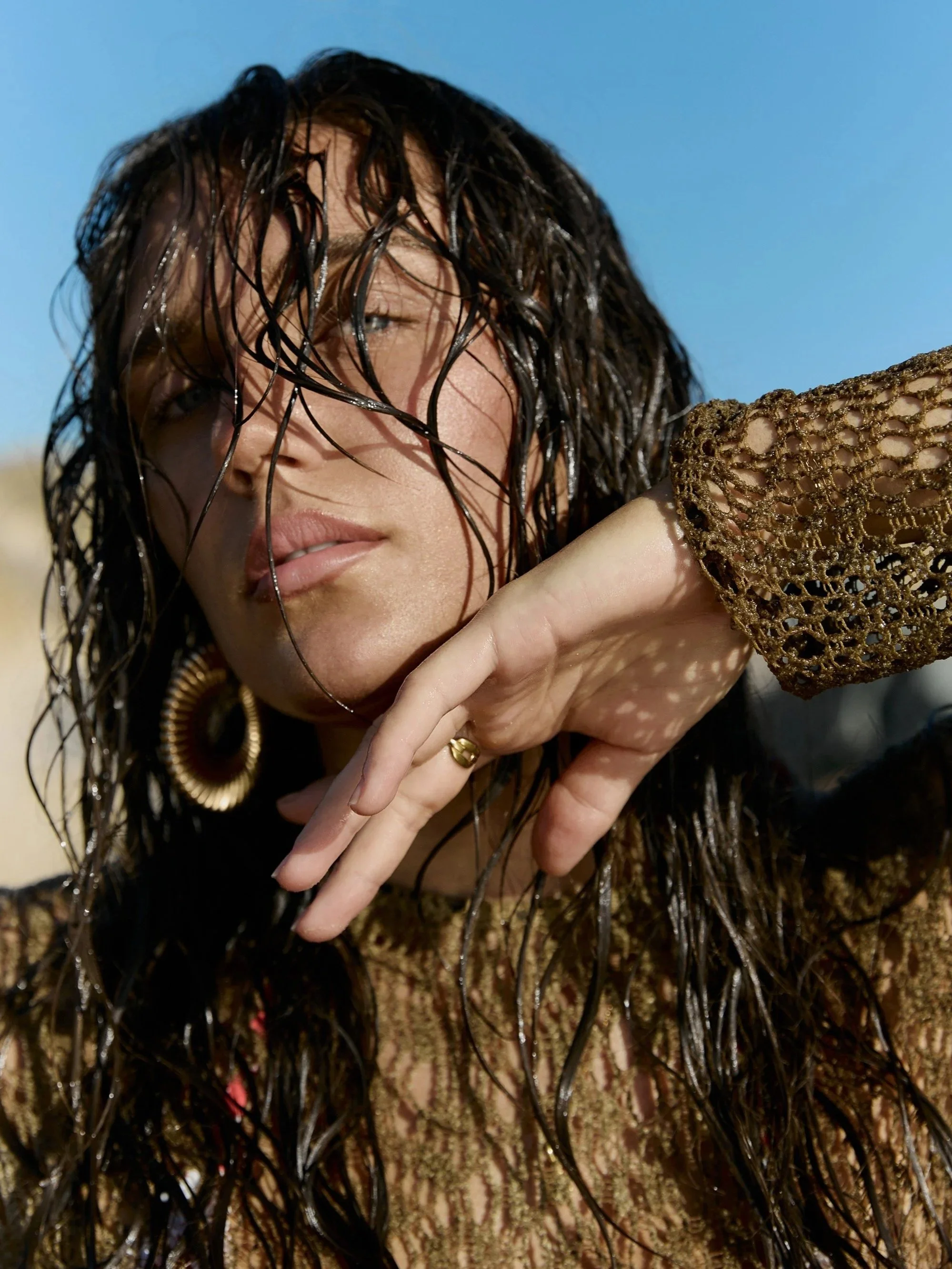 A woman with wet dark hair, wearing large gold earrings and a gold woven top, touching her face against a bright blue sky.