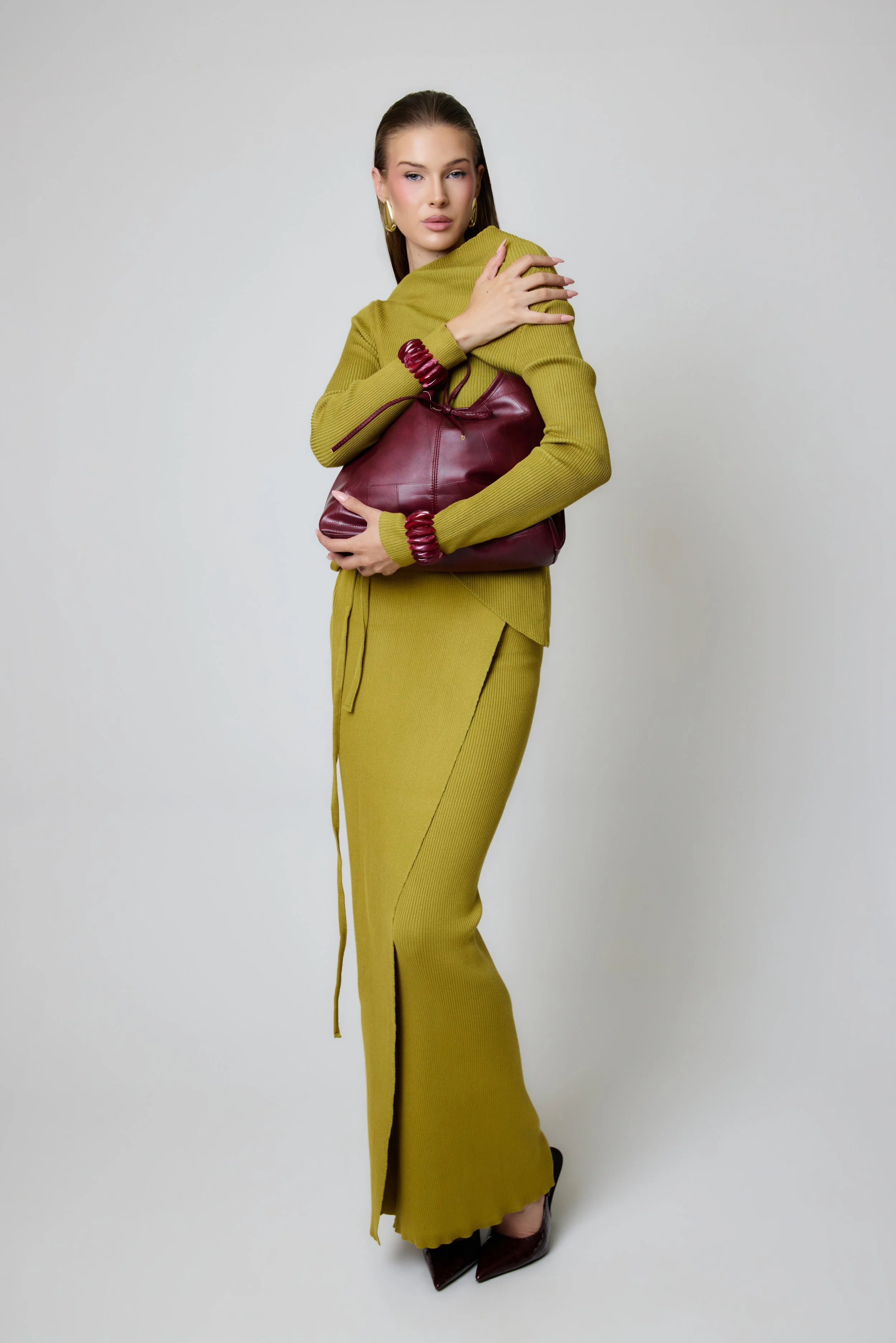 A woman wearing a mustard yellow long dress with a matching cardigan, holding a burgundy leather handbag and wearing matching red bangles on her wrists. She has long brown hair, gold hoop earrings, and is standing against a plain light gray backgroun