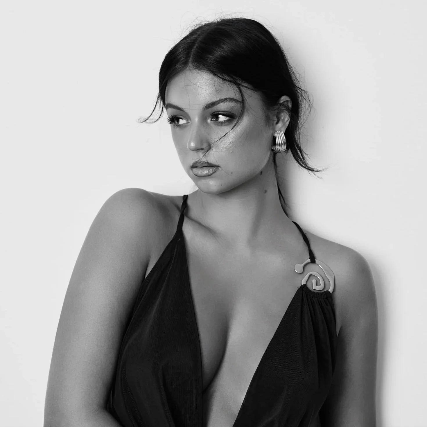 A woman with dark hair styled in a short bob, wearing hoop earrings and a sleeveless, deep V-neck satin dress with a decorative necklace, looking to her right with her arms crossed.