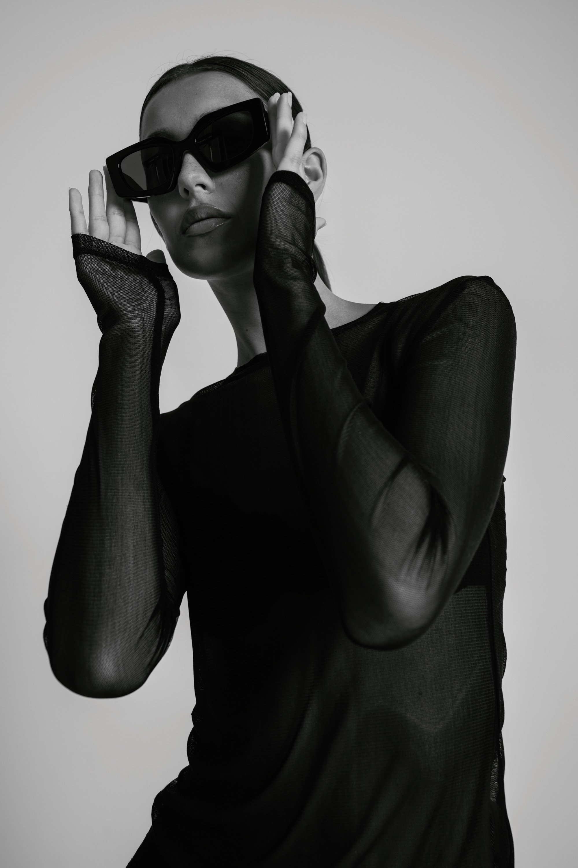 A woman wearing large black sunglasses, a sheer black long-sleeve top, and gloves holds her hands near her ears, with a neutral expression on her face. The background is plain and light-colored.