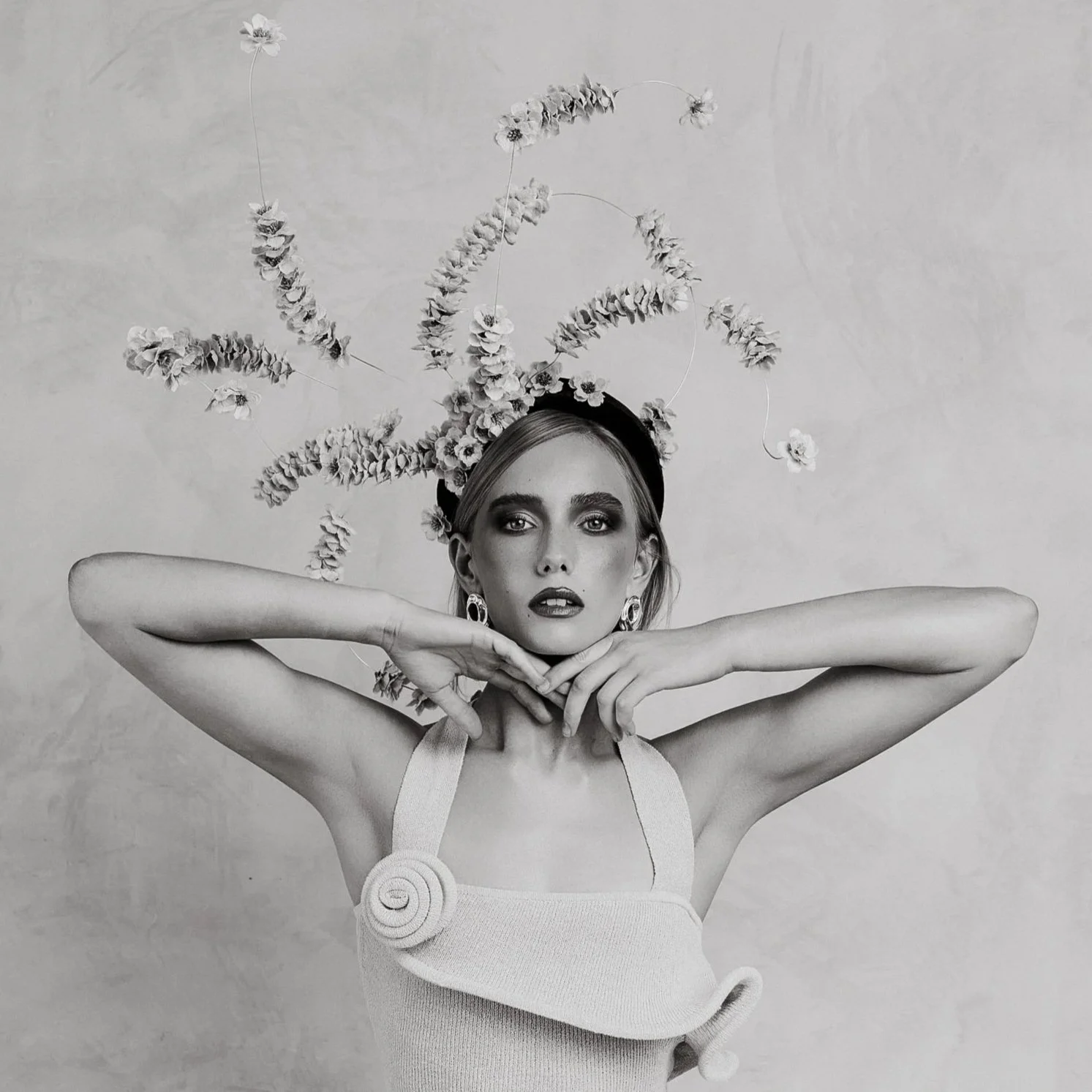 Black and white photo of a woman wearing earrings, a top with shoulder straps, and a headpiece made of flowers and curved wire, posing with her arms raised and forming a triangular shape around her face.