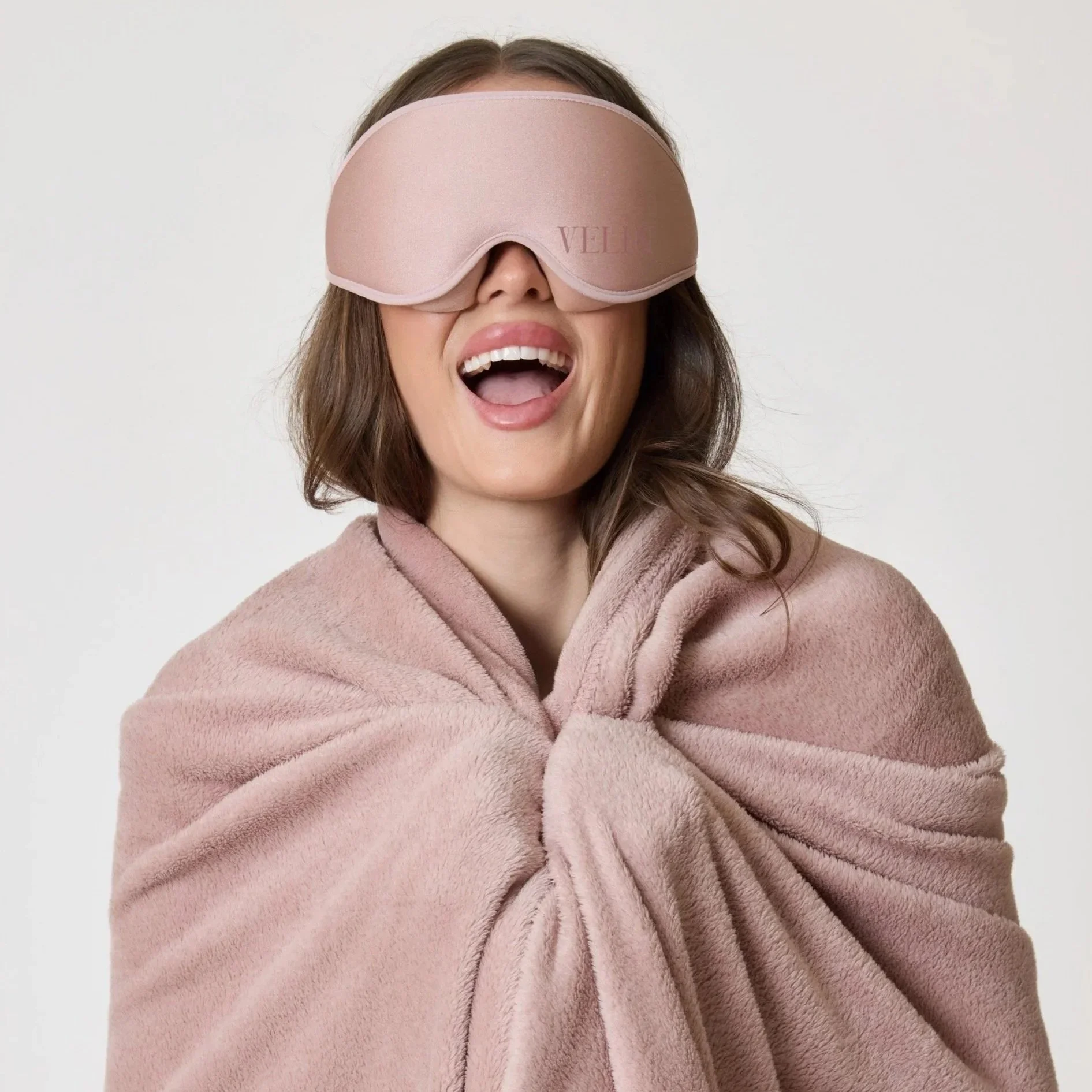 A woman with long brown hair, smiling with her mouth open, wearing a soft pink bathrobe and a pink sleep mask covering her eyes. The background is plain white.