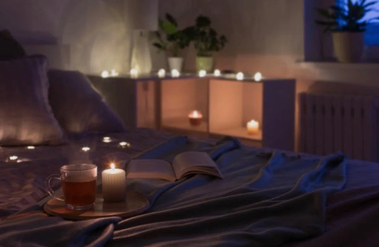 Cozy bedroom with dim lighting, candles, a cup of tea, an open book on the bed, and potted plants on a shelf in the background.