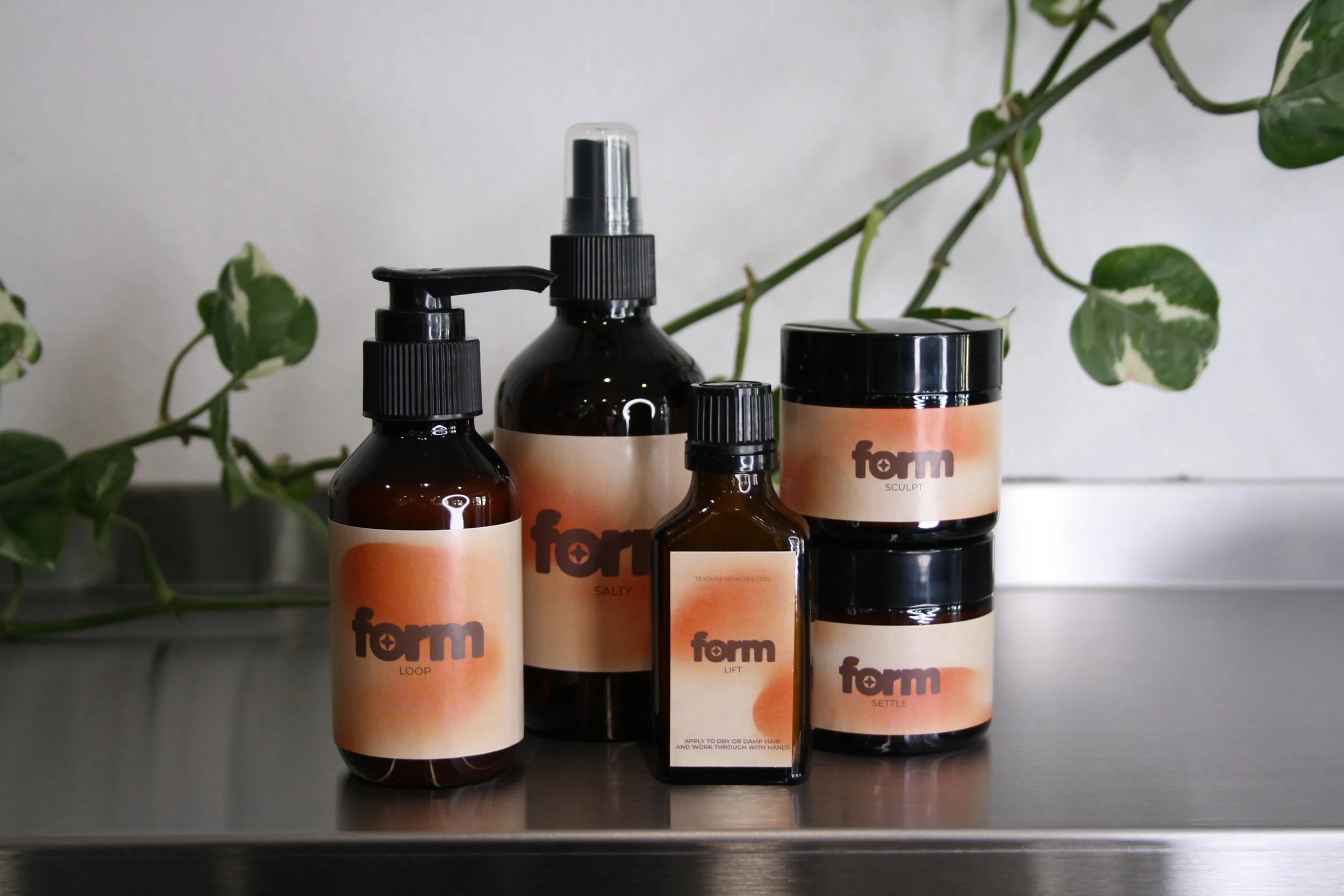 Collection of hair care products in bottles and jars with beige labels and black caps, set on a dark surface with green leafy background.