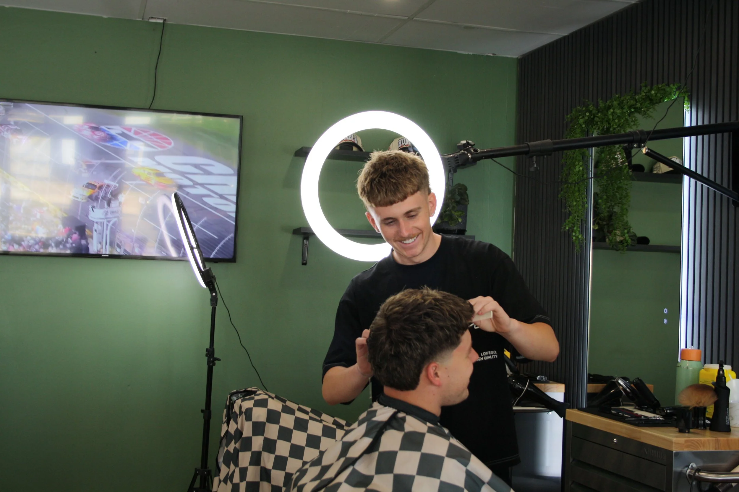 A barber smiling while giving a haircut to a customer with a checkered shirt in a modern salon with green walls, illuminated ring light, and a large monitor showing a race car event.