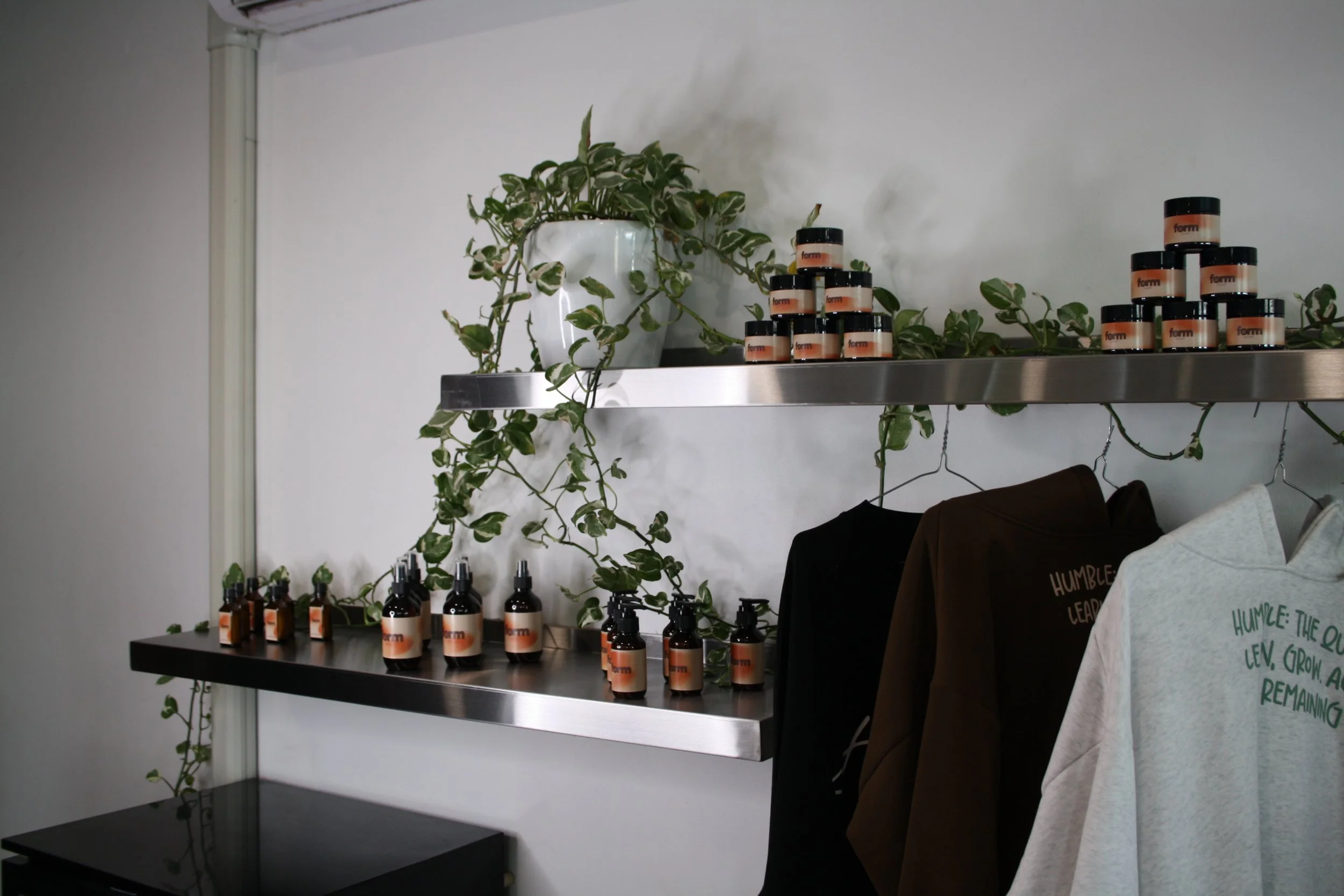 Shelves with skincare or cosmetic products, green plants, clothing hanging on hooks, and bags against a plain wall.