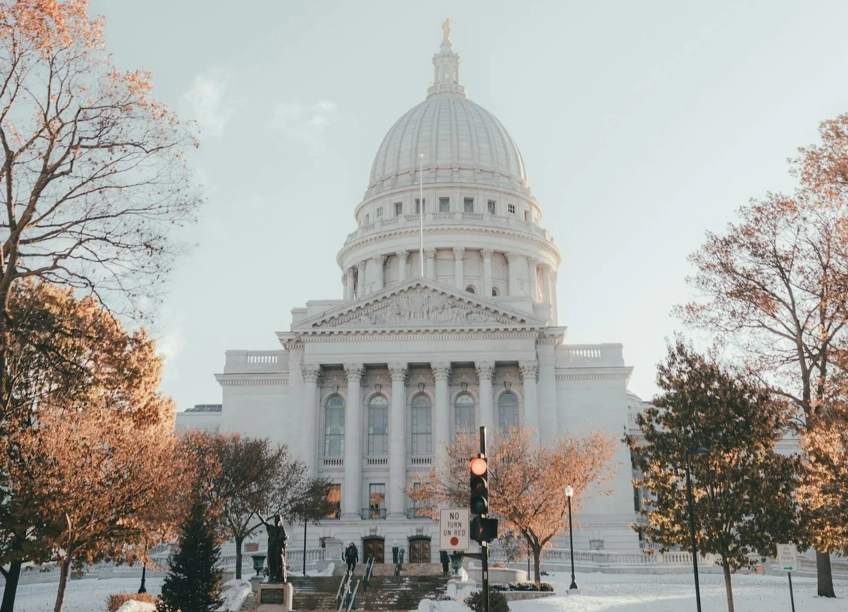 Front view of the U.S. Capitol building with columns, dome, and statue, surrounded by trees with autumn-colored leaves, and snow on the ground.