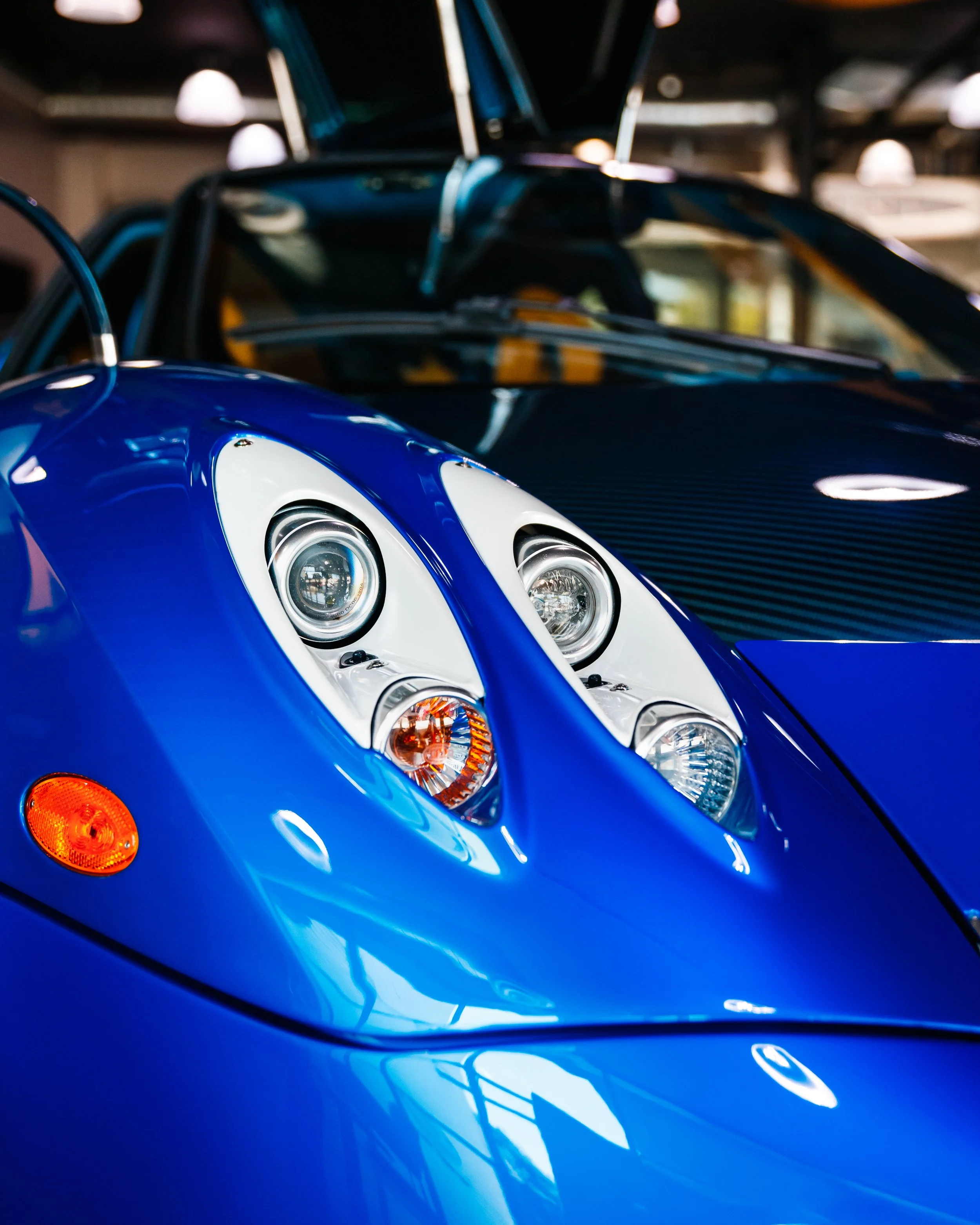 Close-up of a blue Pagani Huayra front with distinctive circular headlights. The background shows an indoor car showroom or garage.