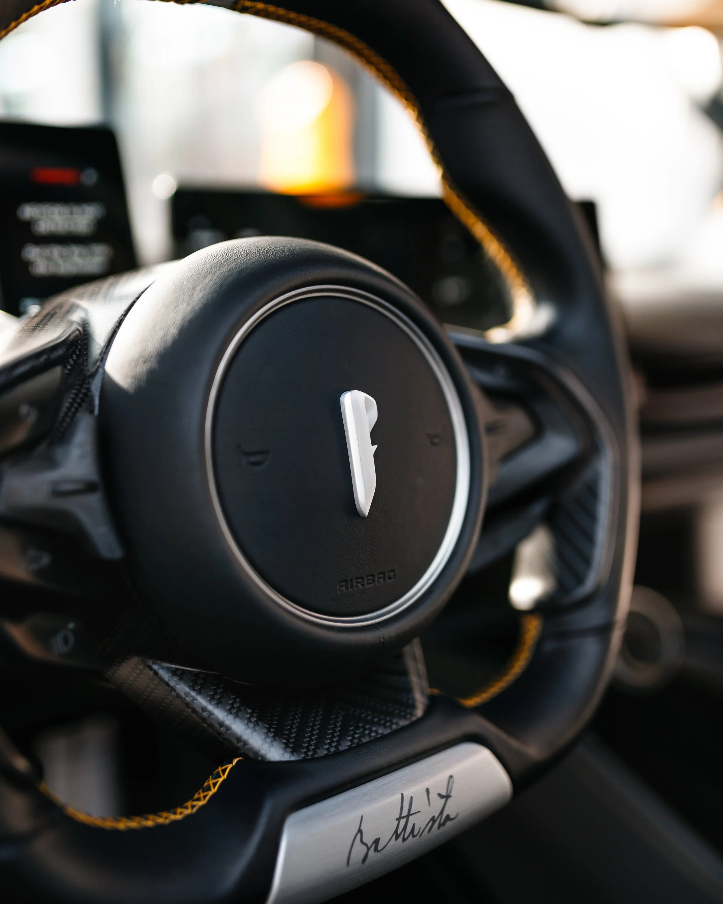 Close-up of a Pininfarina Battista steering wheel with the Pininfarina logo in the center, signature on a metal part at the bottom, and yellow stitching on the rim.