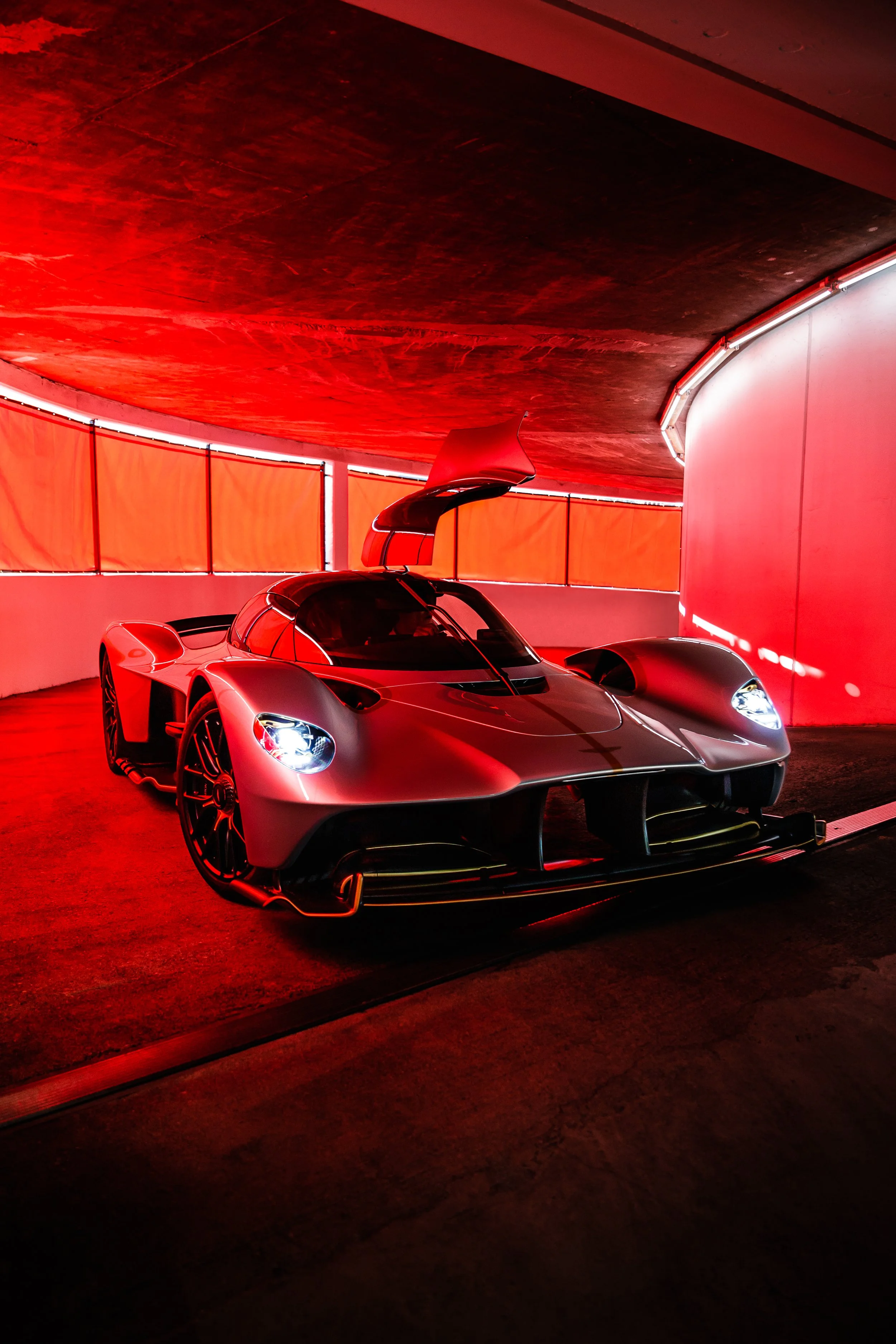 A sleek, silver Aston Martin Valkyrie with its door raised, parked in a red-lit parking garage.