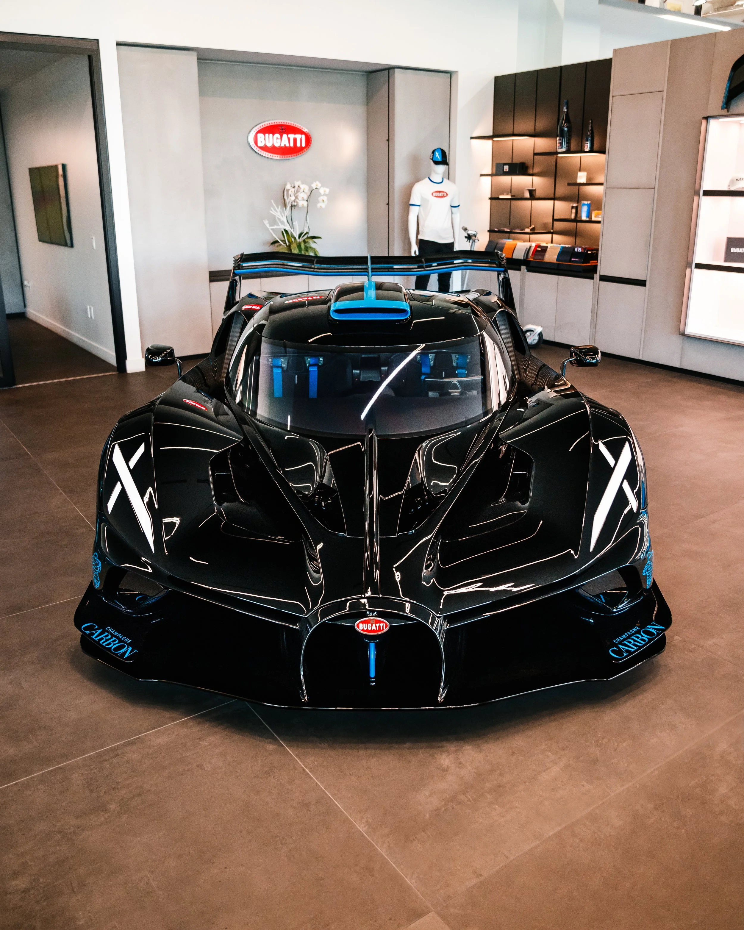 A black Bugatti Bolide displayed indoors with a blue rear wing, in a showroom with automotive memorabilia and a mannequin wearing a Bugatti shirt and cap visible in the background.