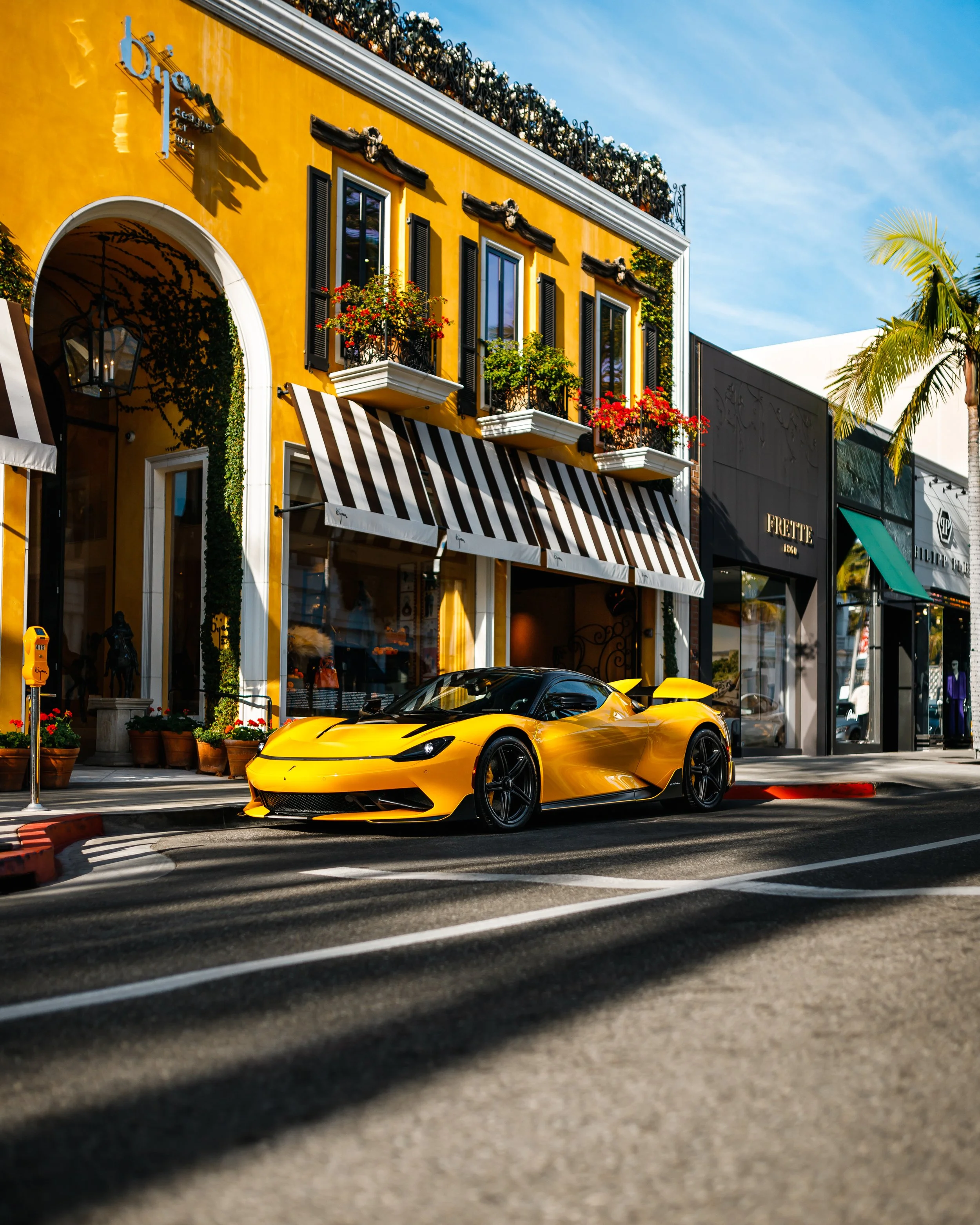 A yellow Pininfarina Battista parked on a city street in front of colorful shops with striped awnings, in a sunny, urban setting with a blue sky and palm trees.