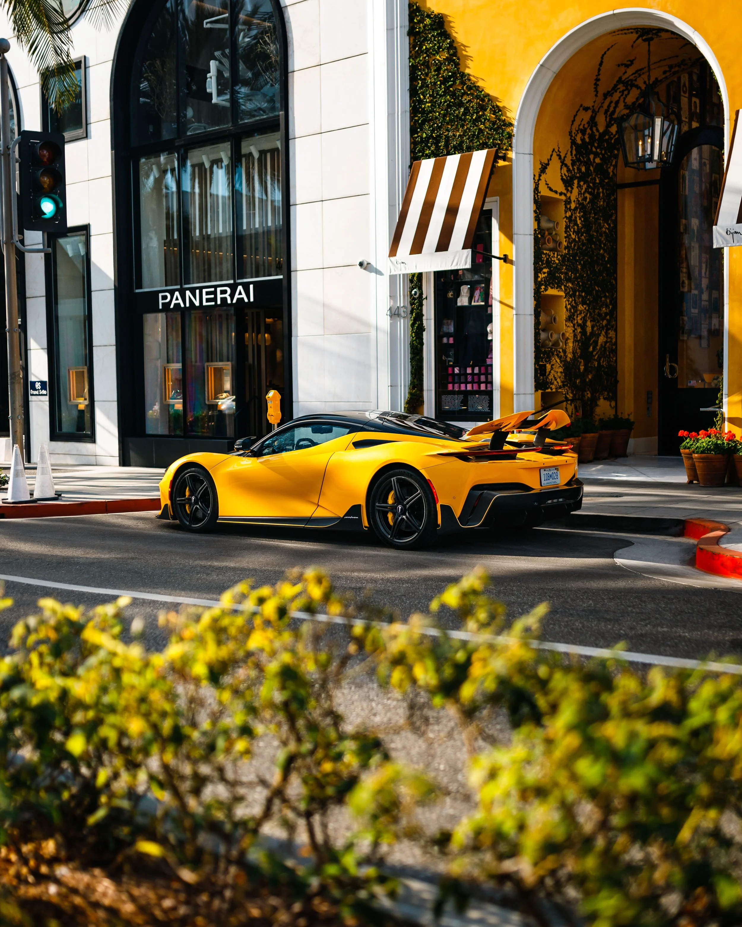 A yellow Pininfarina Battista parked on a city street in front of a storefront with large black-framed windows and a yellow building with a white archway, awnings, and plants.