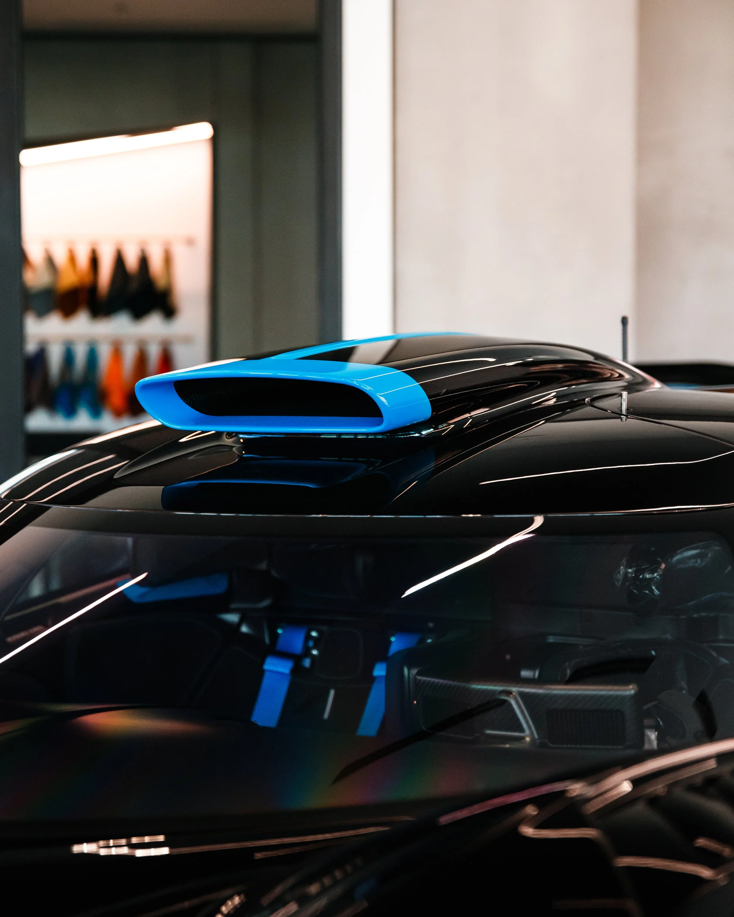 Close-up of a black Bugatti Bolide with a blue air intake on the roof, parked indoors with colorful fabric samples in the background.