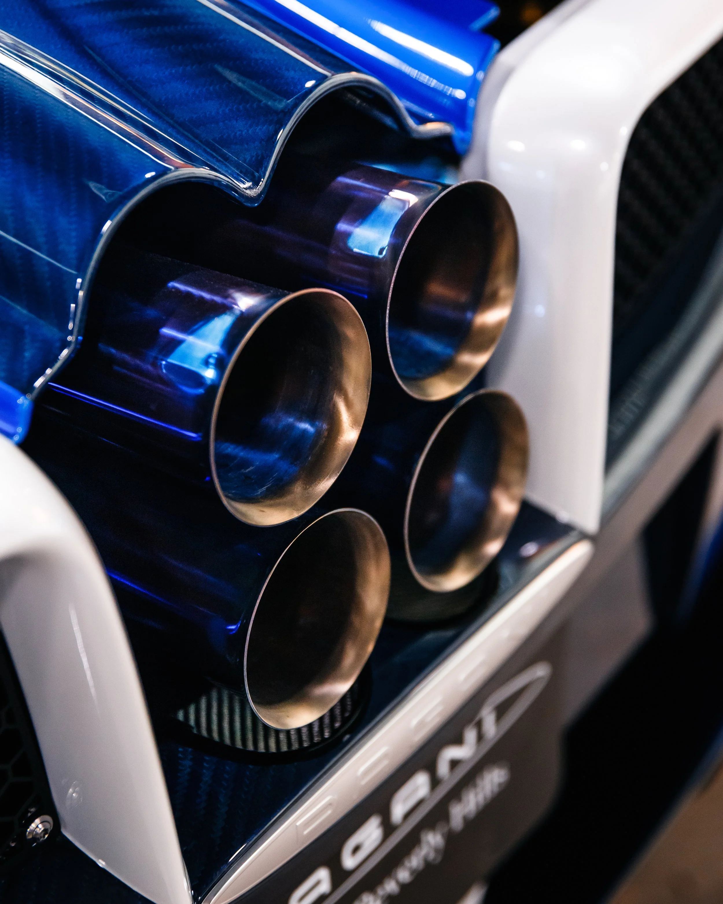 Close-up of a Pagani Huayra's exhaust pipes and rear part, featuring four large, shiny metal exhaust tips against a carbon fiber body and white trim.