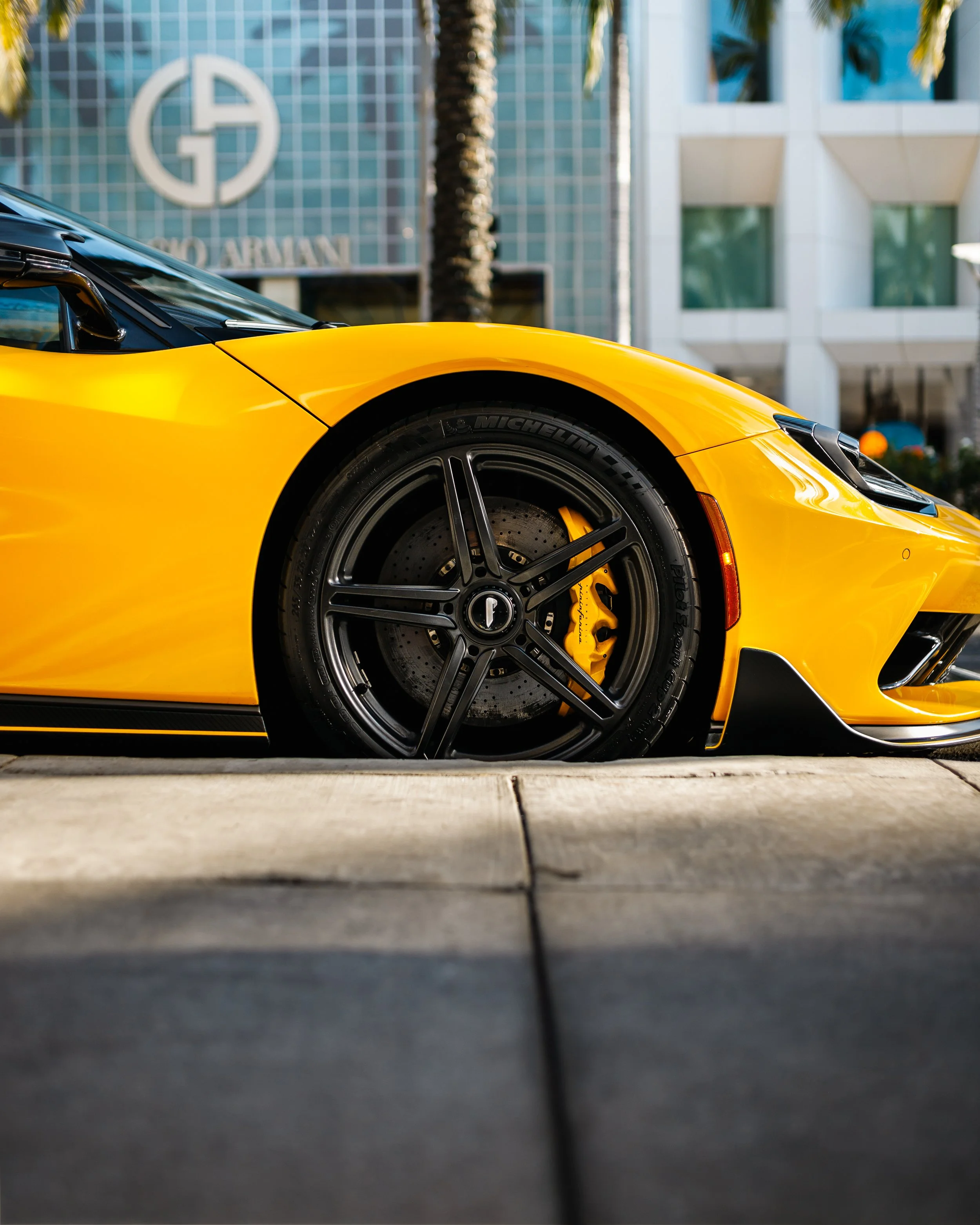 Close-up of a Pininfarina Battista car's front wheel with black rims and brake calipers parked on the street with modern buildings and palm trees in the background.