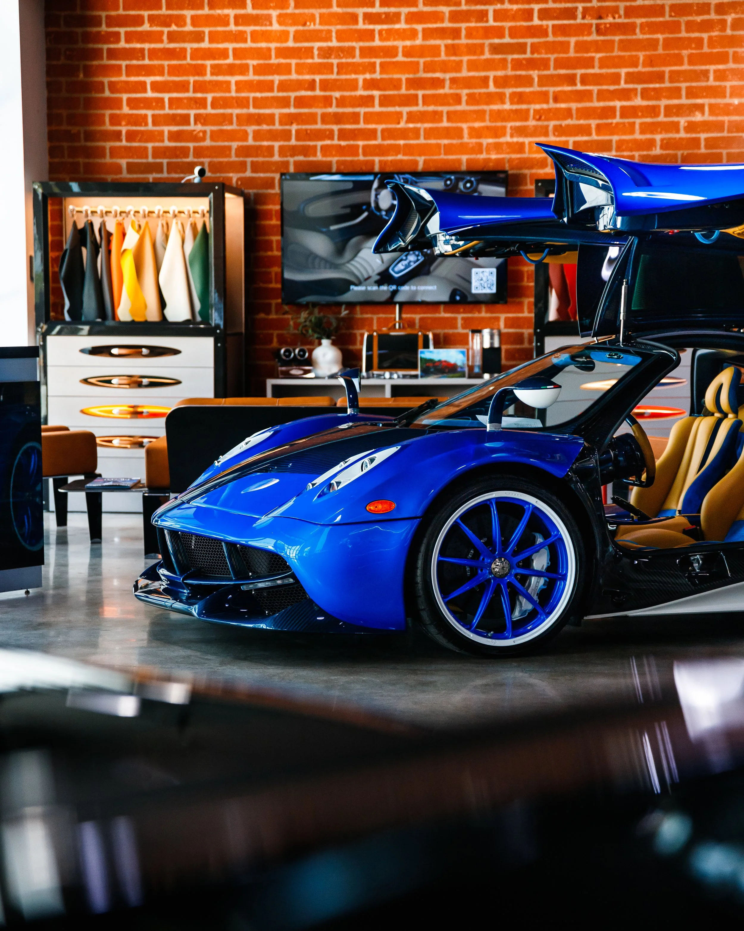 A blue Pagani Huayra with gullwing doors open inside a modern showroom with brick walls and contemporary furniture.