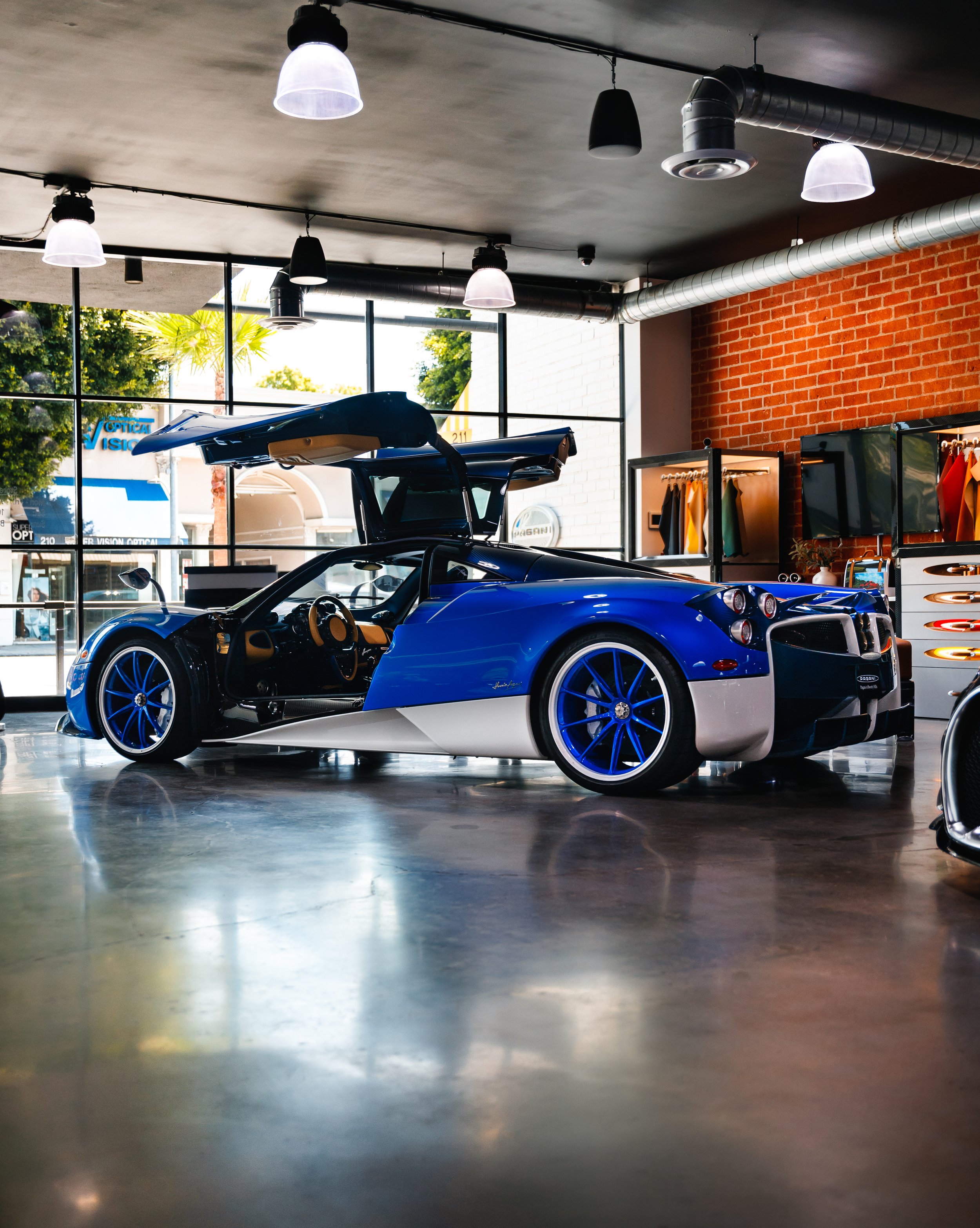 Blue and white Pagani Huayra inside a modern showroom with large windows, brick wall, and clothing display.