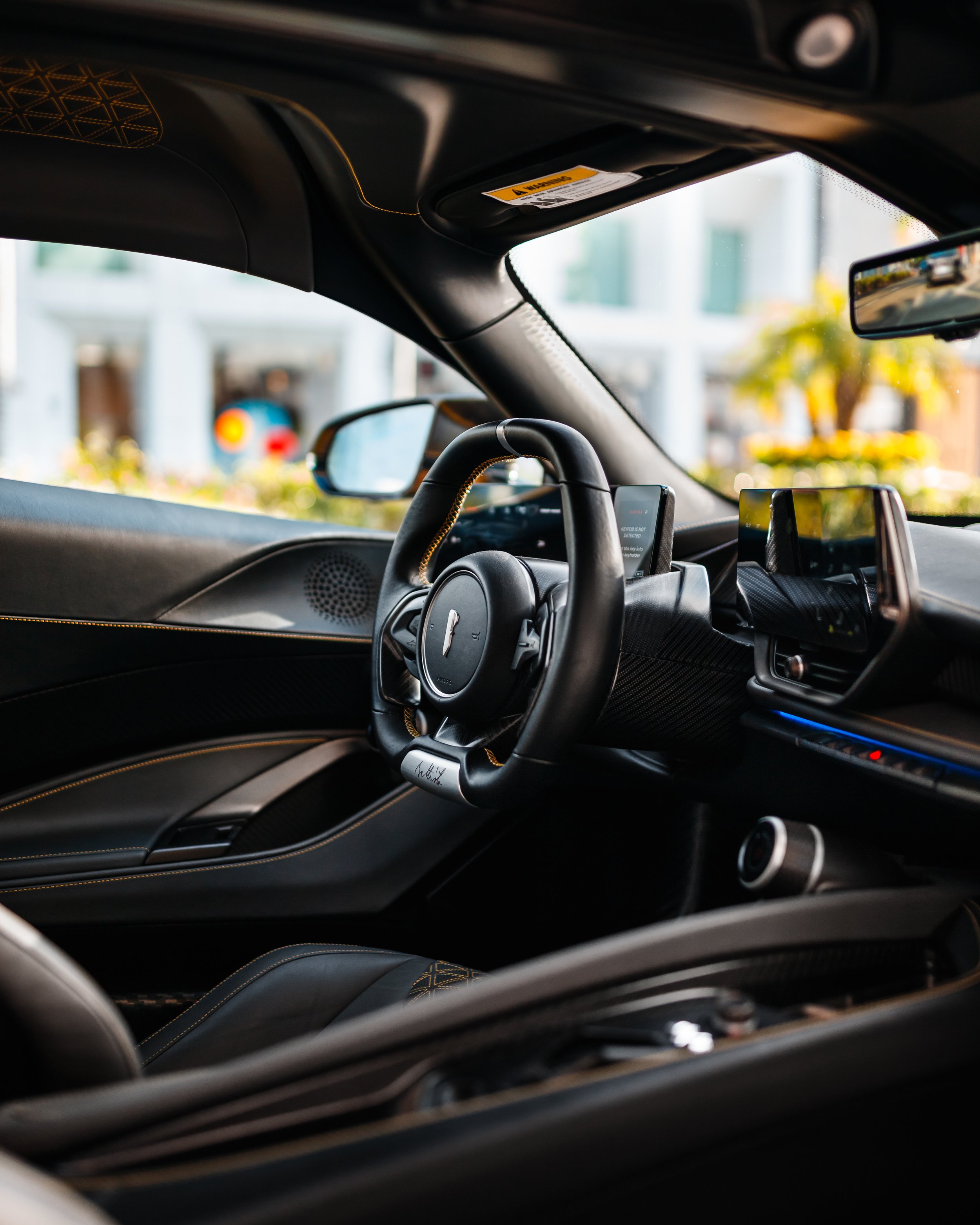 Interior of a Pininfarina Battista showing the steering wheel, digital display screens, and dashboard with a blurred outdoor background.