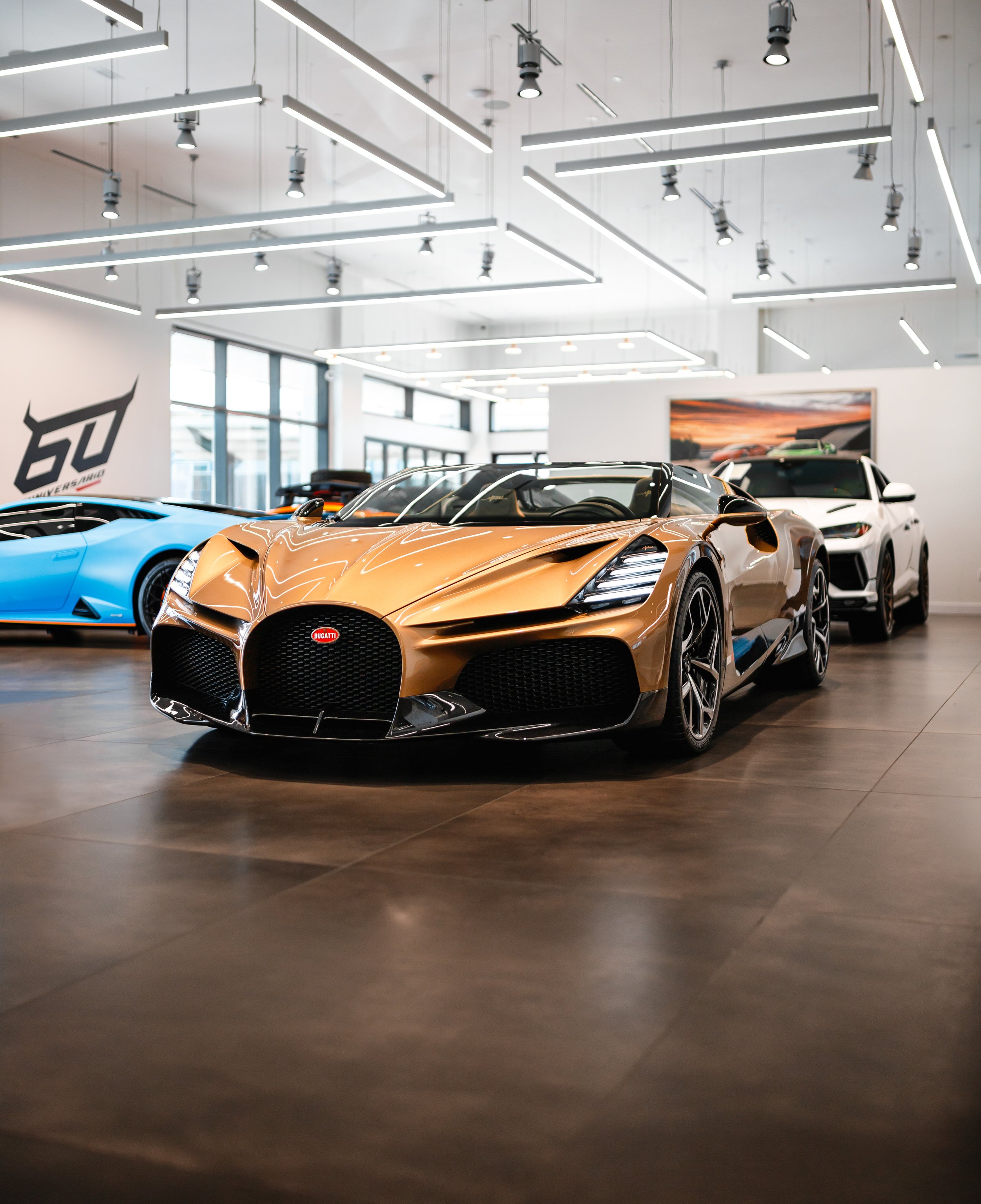 A bronze-colored Bugatti Mistral in a showroom, surrounded by other luxury cars, with bright lighting and modern interior design.