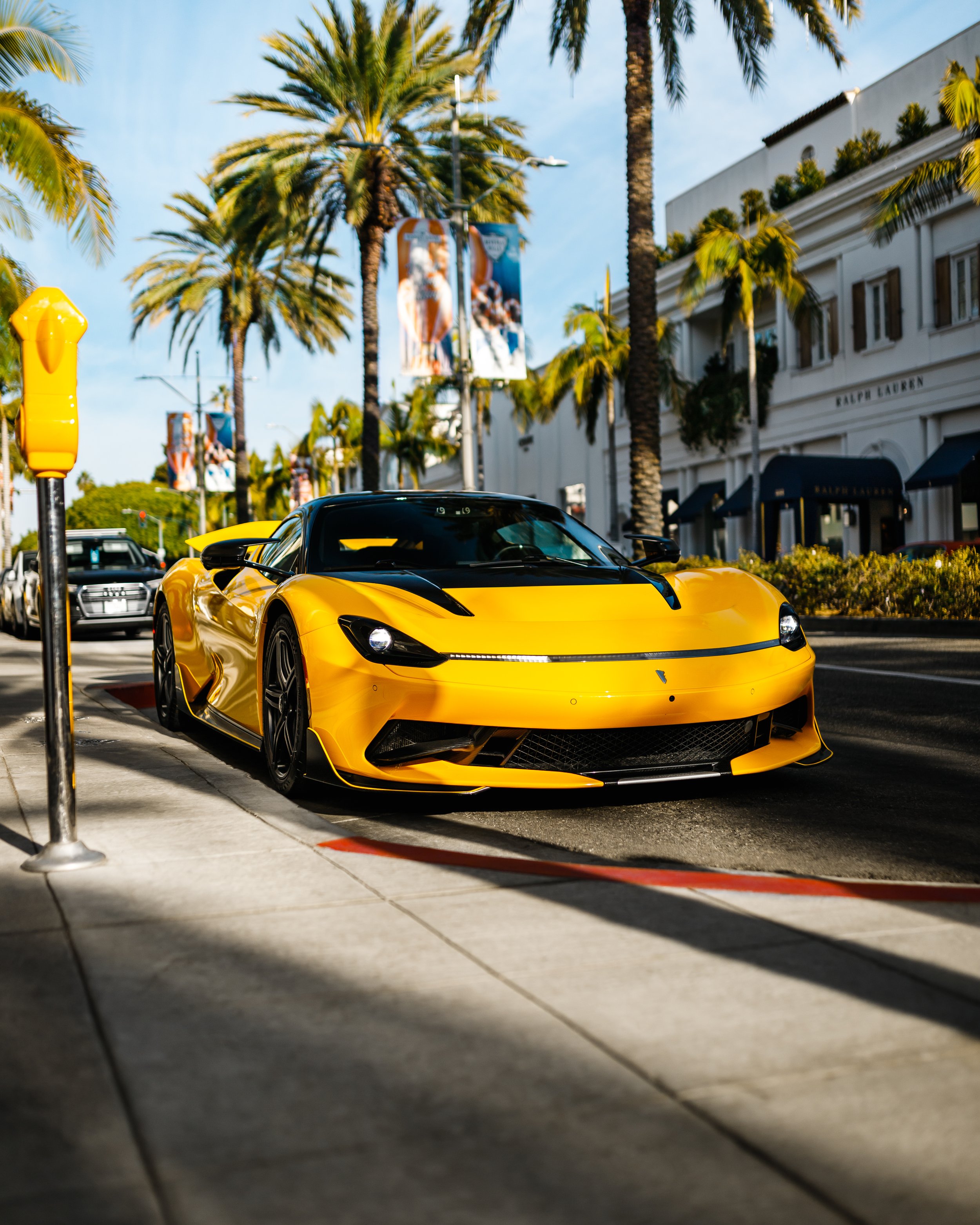 A yellow Pininfarina Battista parked on a street lined with palm trees and high-end shops.